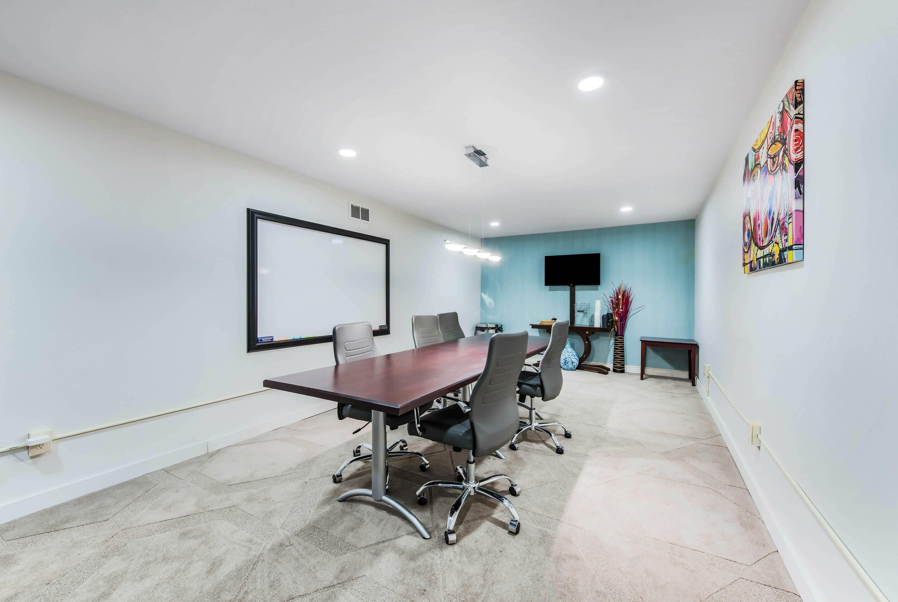 A conference room with a long table, four chairs, a projector screen, and a flat-screen TV.