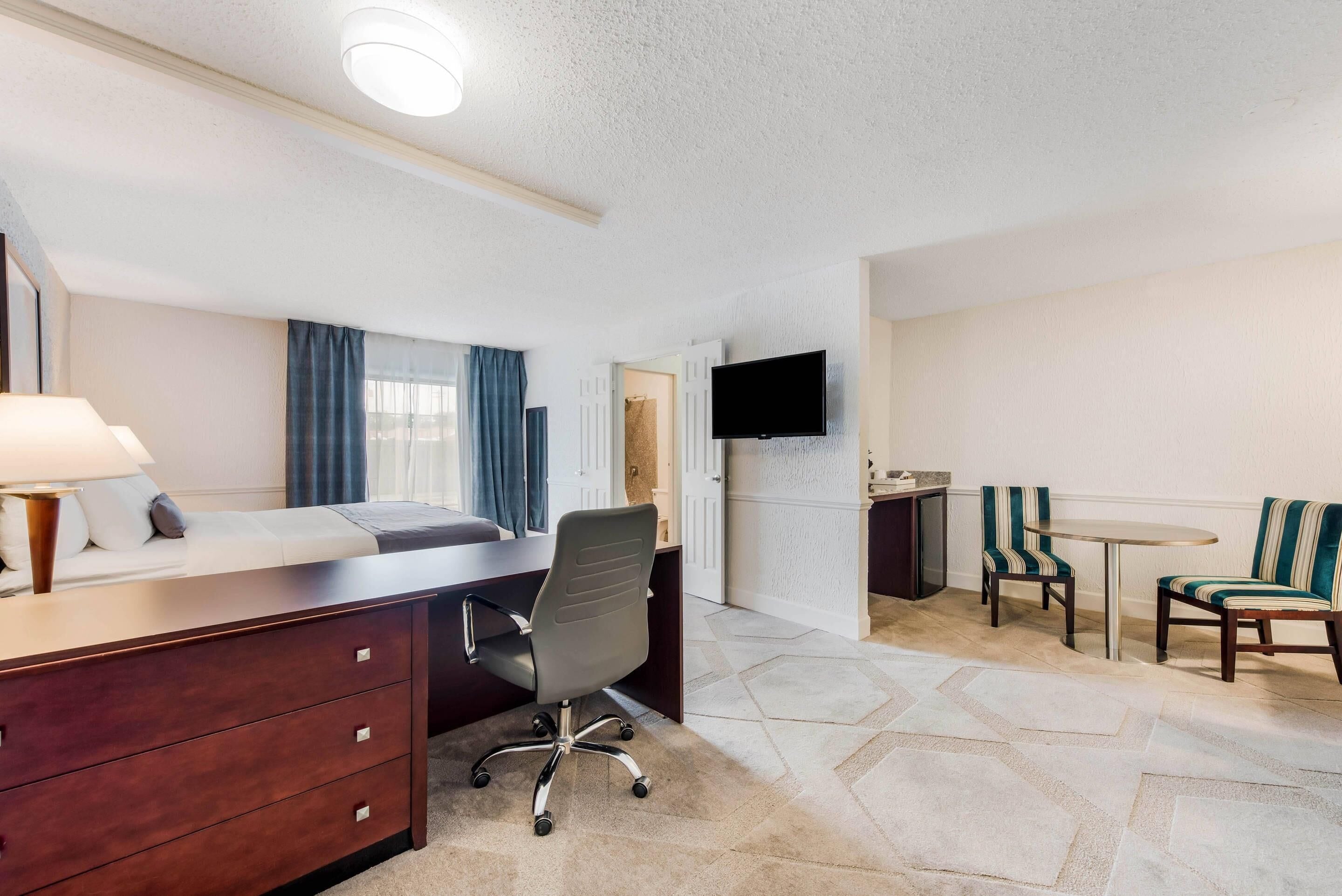 A hotel room with a bed, desk, office chair, TV, and two striped chairs.