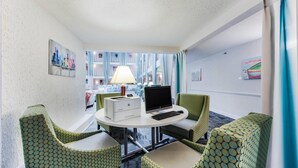 A hotel room with a round table, two green patterned chairs, a desk with a computer and printer, a lamp, and a view of a building with balconies.