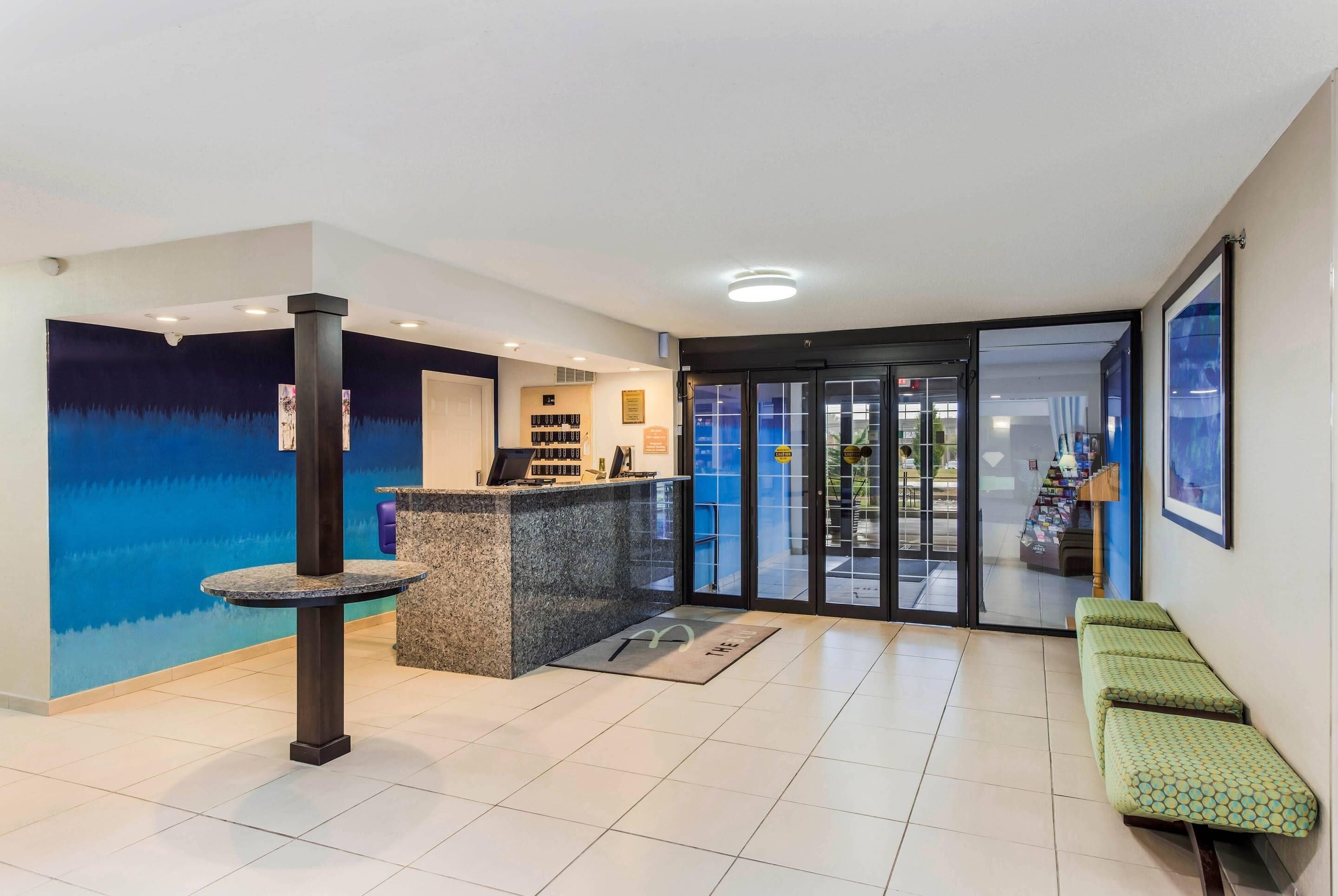 Ein Hotel-Empfangsbereich mit Empfangsschalter, einer Sitzecke mit grünen Polsterbänken und einer Glastür zum Außenbereich.