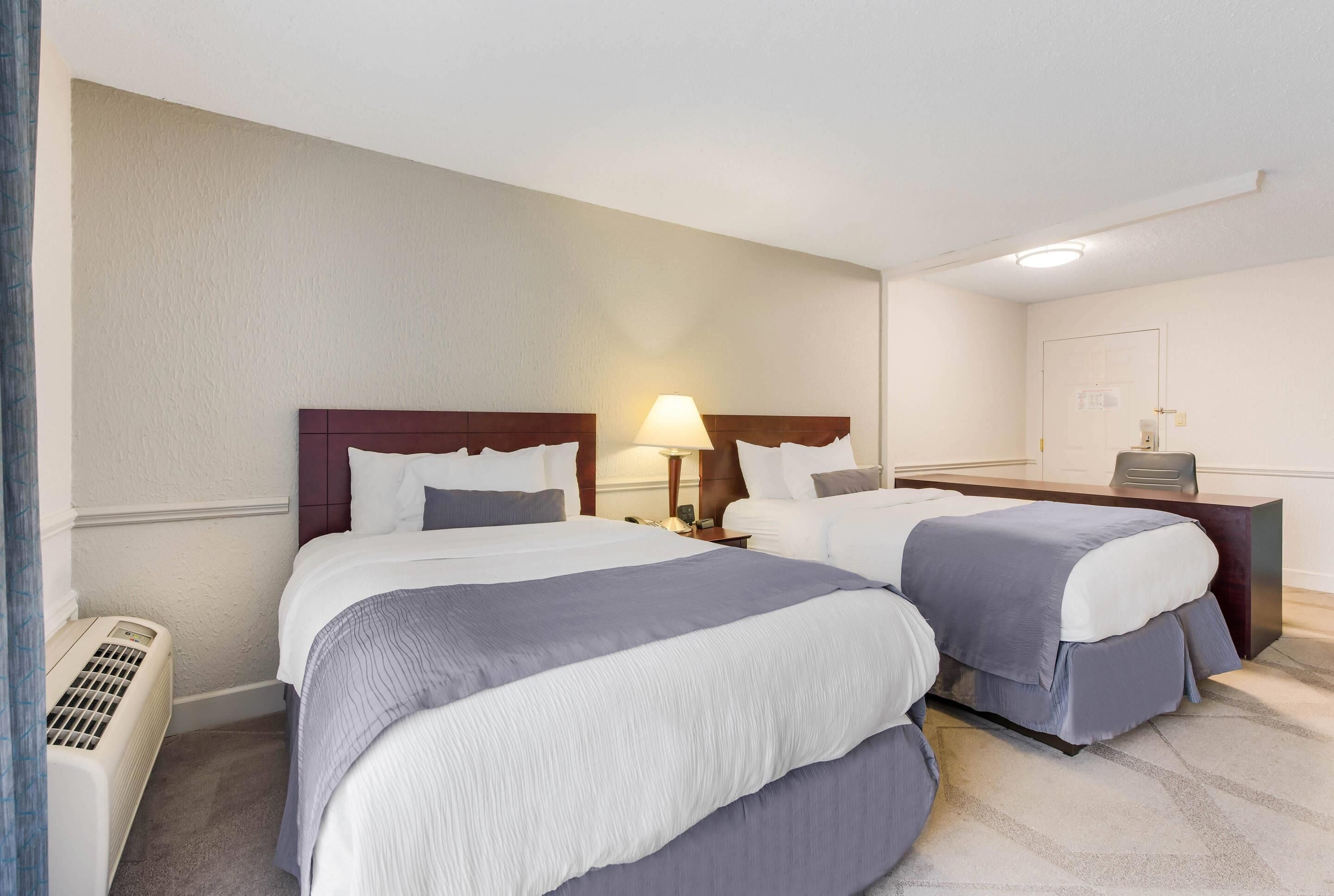 Habitación de hotel con dos camas, una mesita de noche con lámpara, un calentador y un pequeño escritorio con silla.