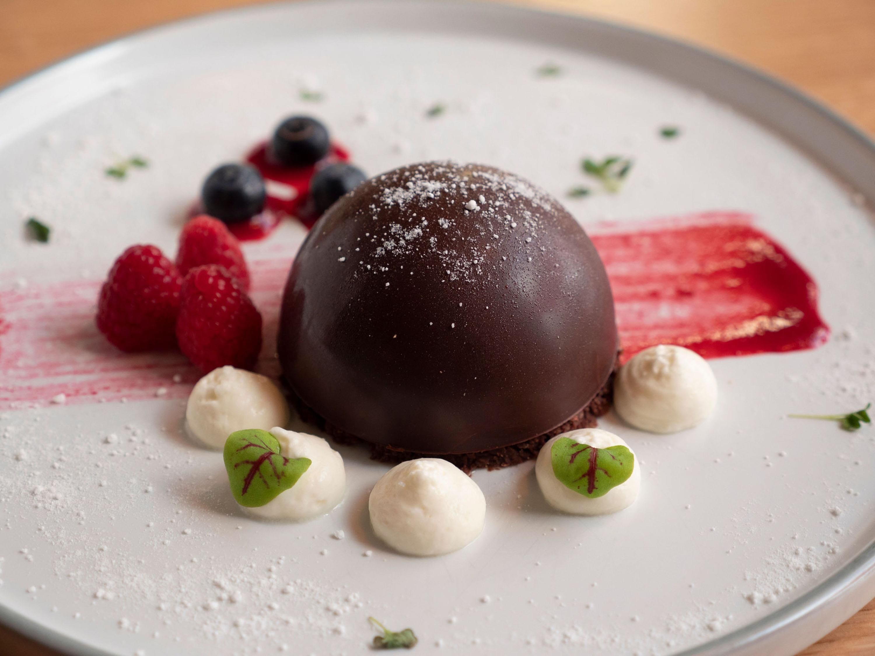 A dessert plate with a chocolate sphere, raspberries, blueberries, and small heart-shaped items.