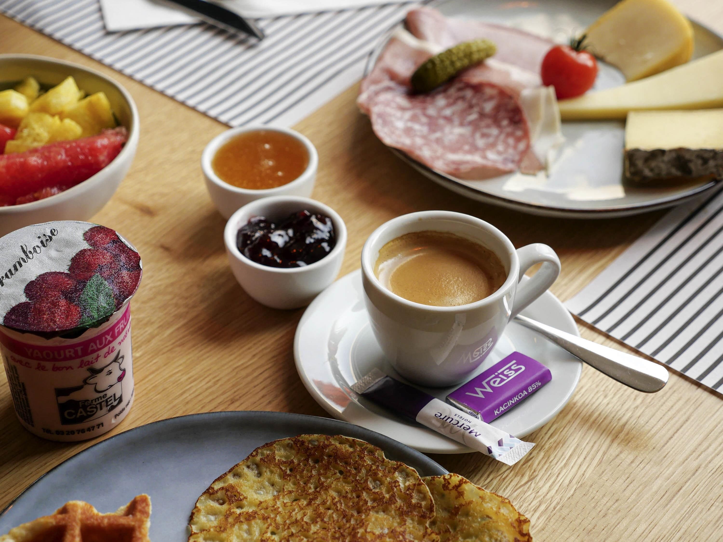 Een ontbijttafel met een bord vleeswaren, kaas en fruit, een kop koffie en een potje jam.