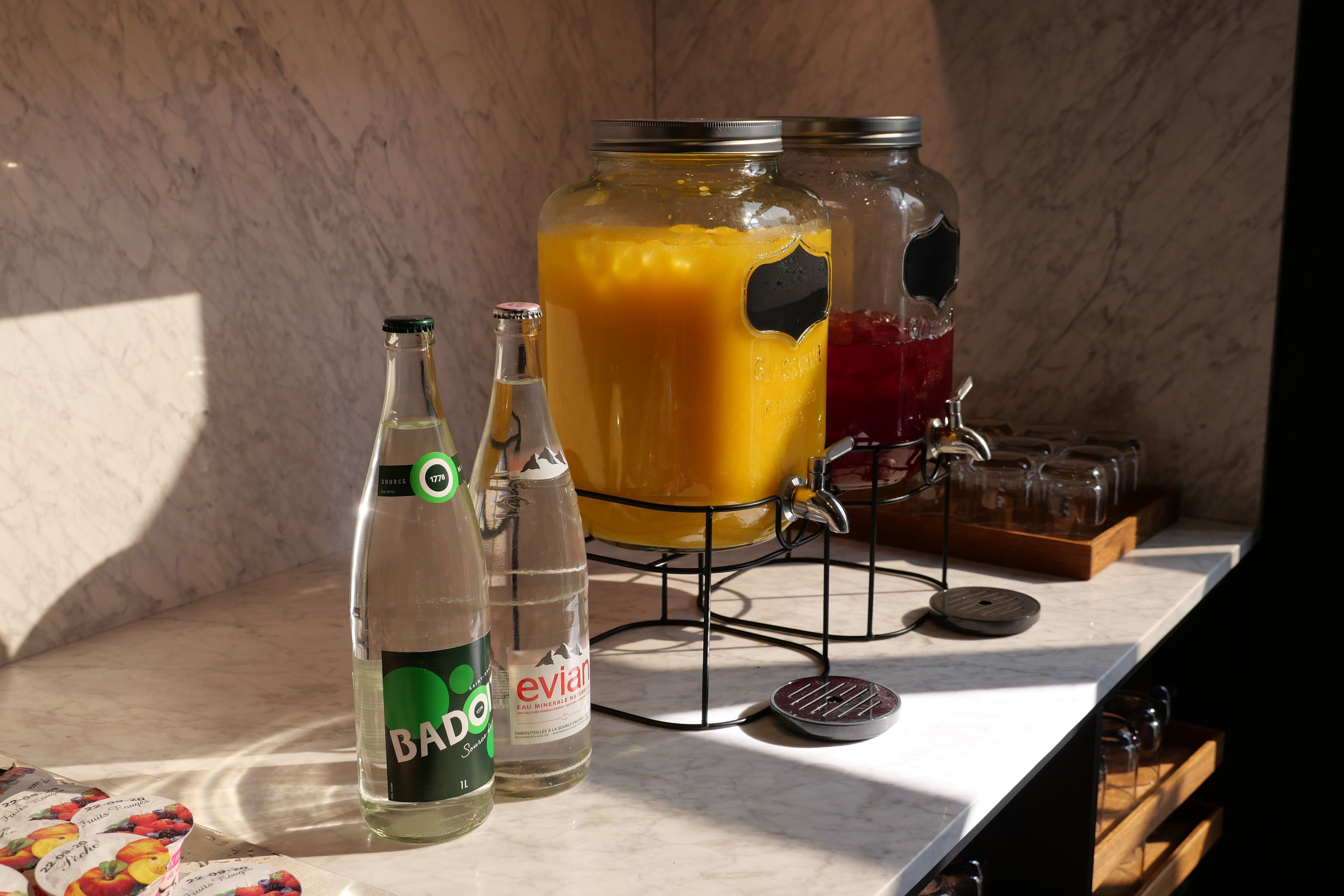 En köksbänk med en glasburk fylld med en gul vätska, två flaskor märkta med "Bado" och "Evian", och ett metallställ som håller glasburkar.