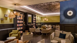 A modern lounge area with a variety of seating options, including sofas and chairs, arranged around a central table. There are bookshelves filled with books and decorative items, and a large wall with a circular design element. - Mercure Lille Marcq en Baroeul (Marcq-en-Baroeul)