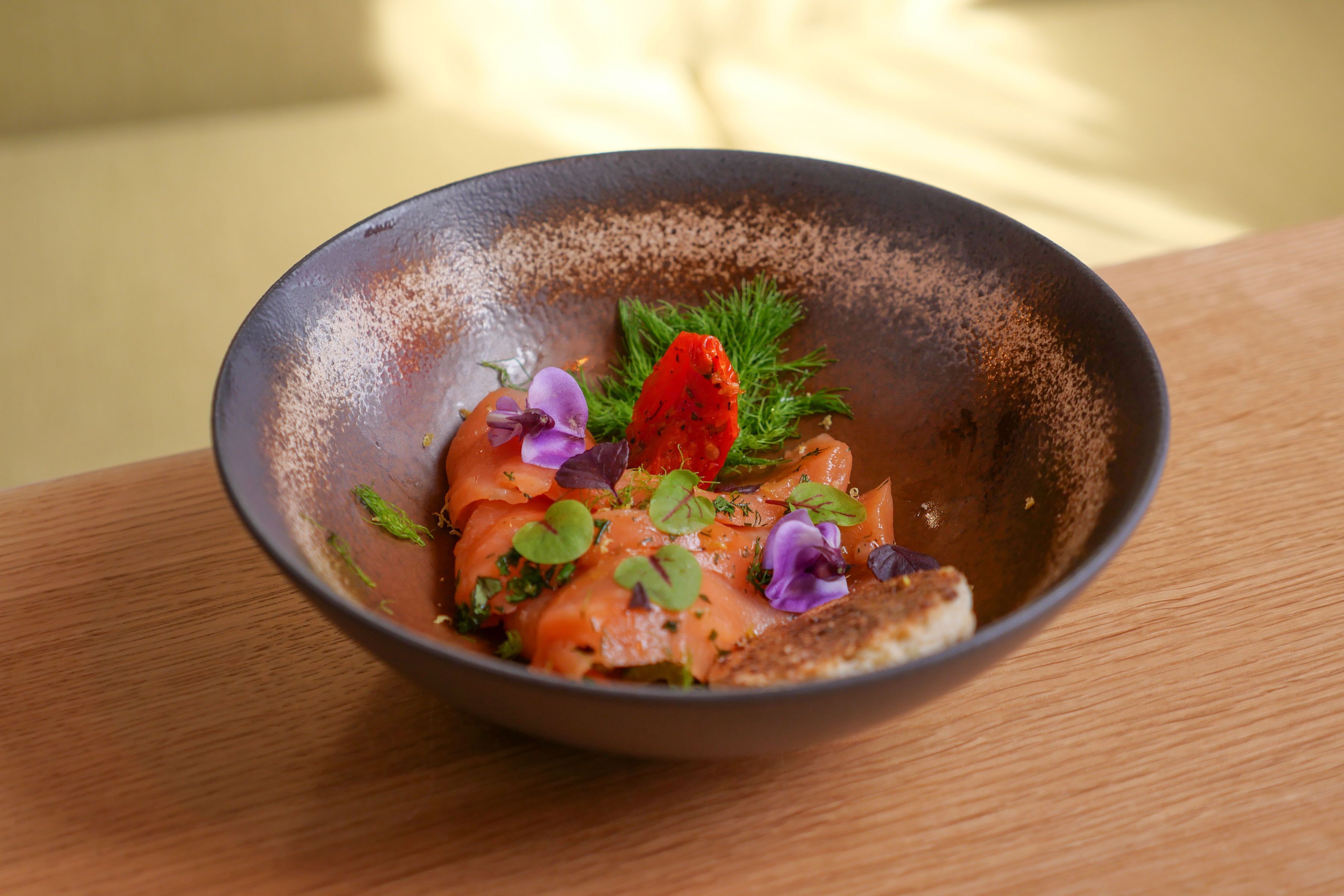 Eine Schüssel mit Essen, darunter Lachs, grünes Gemüse und eine rote Dekoration, auf einem Holztisch.