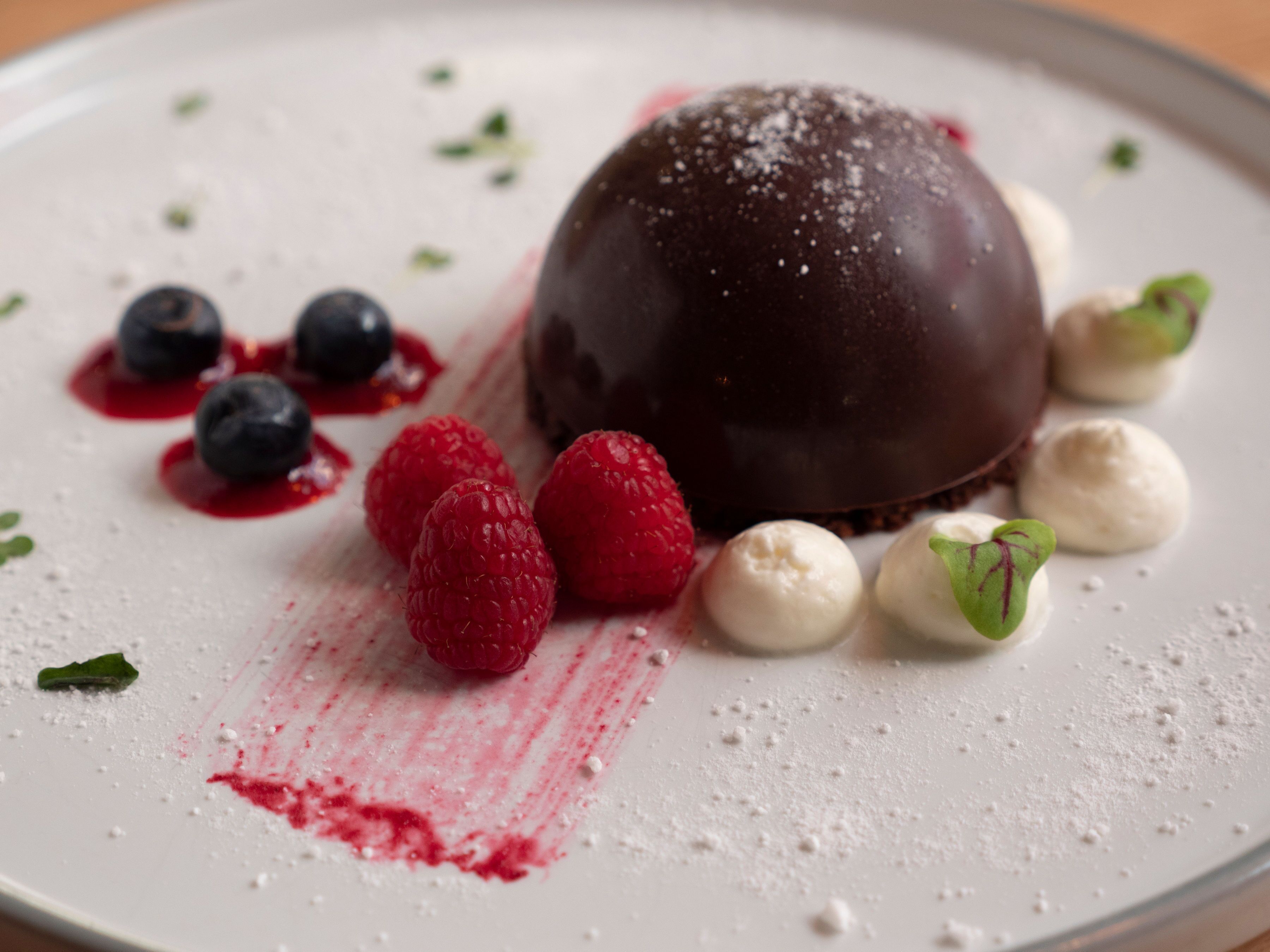 A dessert plate with a chocolate sphere, raspberries, blueberries, and a dusting of powdered sugar.