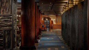 A corridor with a patterned carpet, wooden columns, and a bar area in the distance. - Royalton New York (New York)