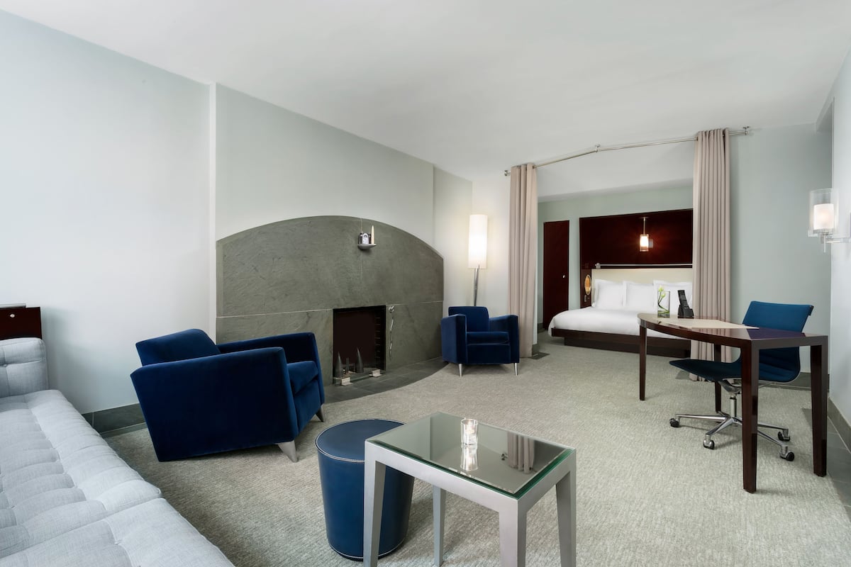 Quarto de hotel com sofá, duas poltronas, mesa de centro de vidro, um pequeno banquinho, lareira, cama, escrivaninha e abajur.