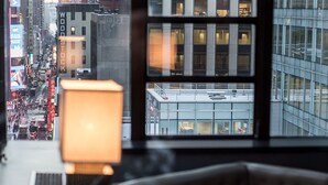 Vista desde una ventana que muestra un paisaje urbano con un edificio moderno, una calle con vehículos y una lámpara sobre una mesa.
