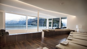 Ein moderner Wellnessraum mit großem Fenster mit Blick auf einen Bergsee, Holzboden und einer Sitzecke mit Handtüchern.