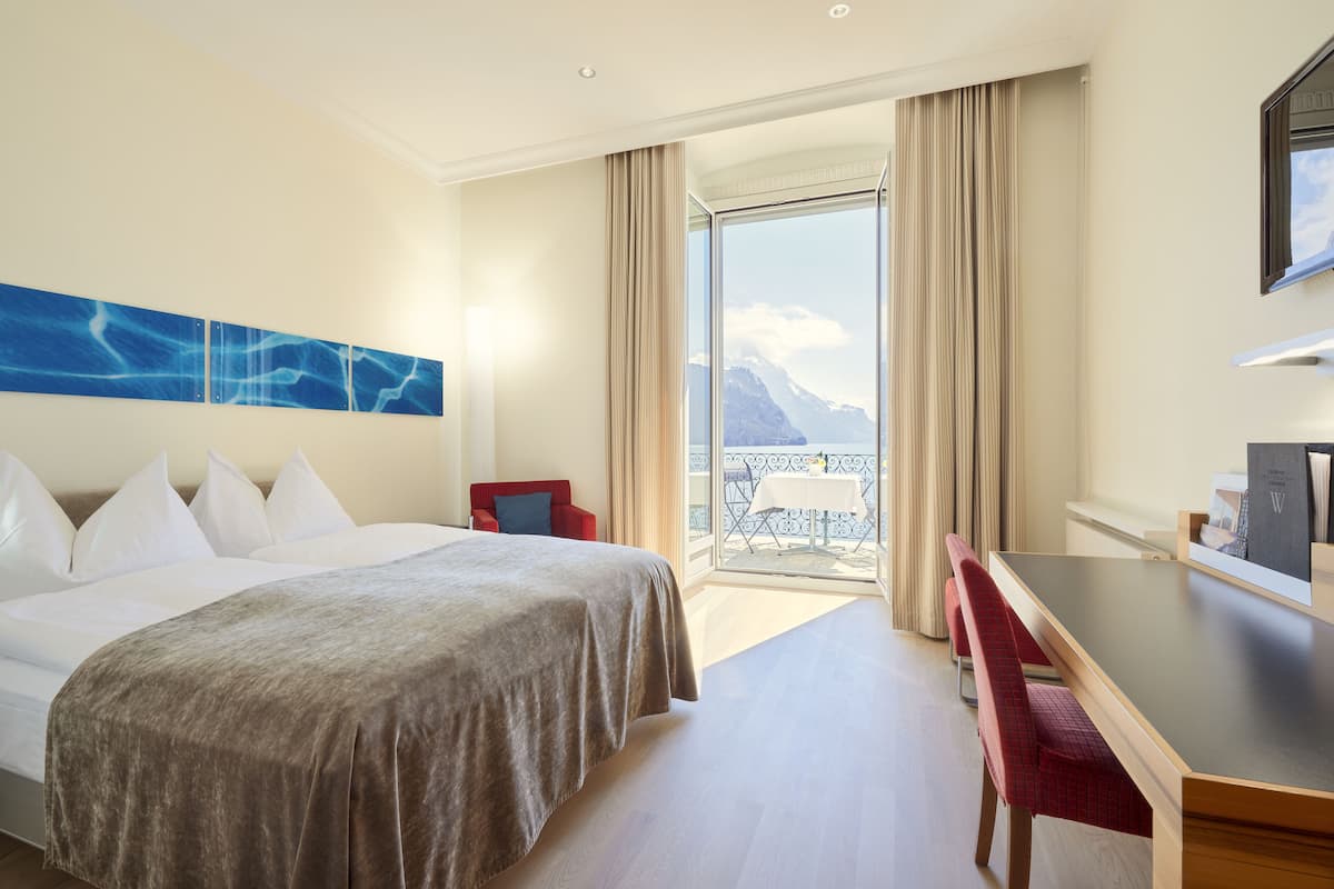 Une chambre d’hôtel avec un lit, un bureau, une chaise, une vue sur les montagnes depuis un balcon et un tableau au mur.