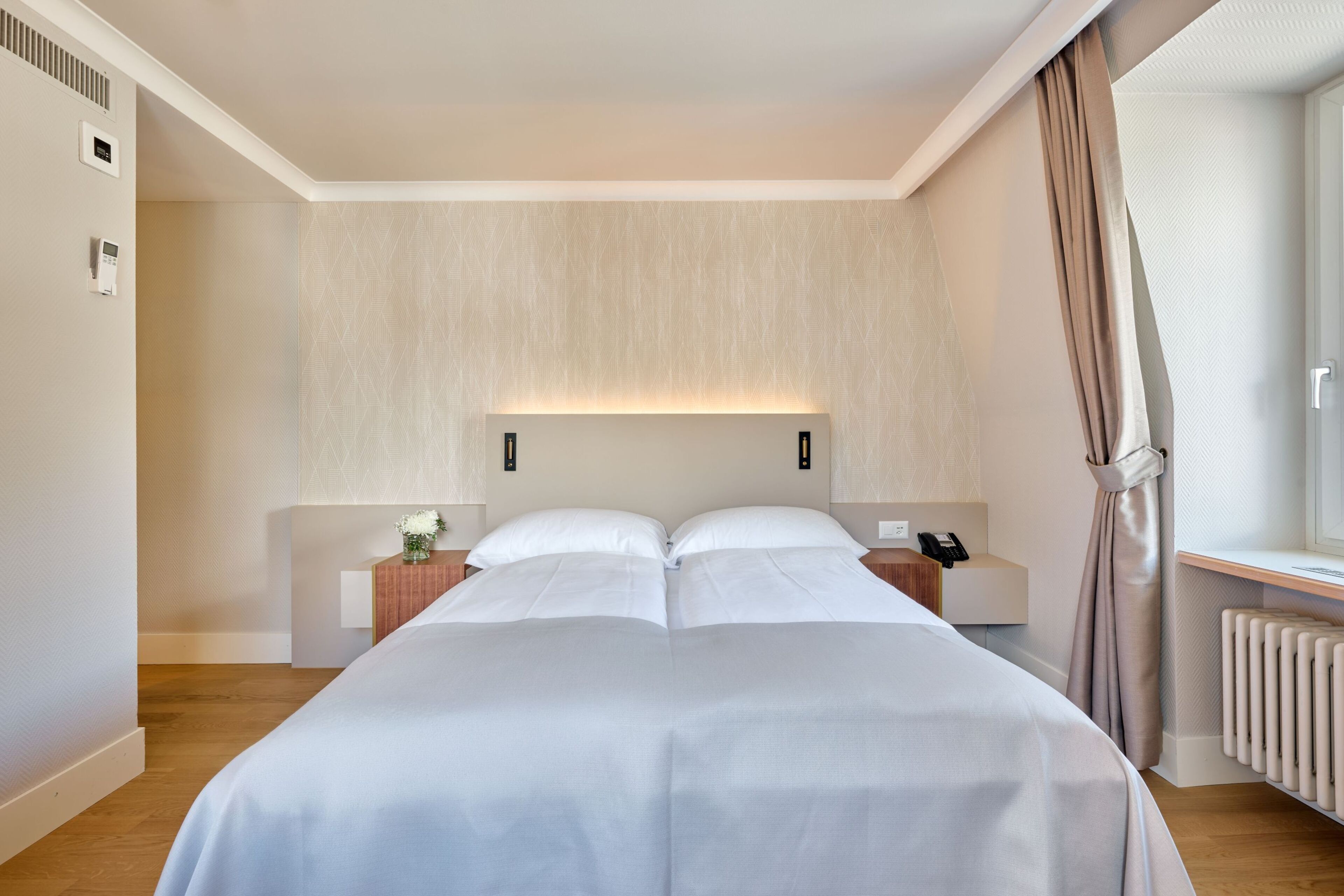Habitación de hotel con una cama grande, mesitas de noche, termostato en la pared y una ventana con cortinas.