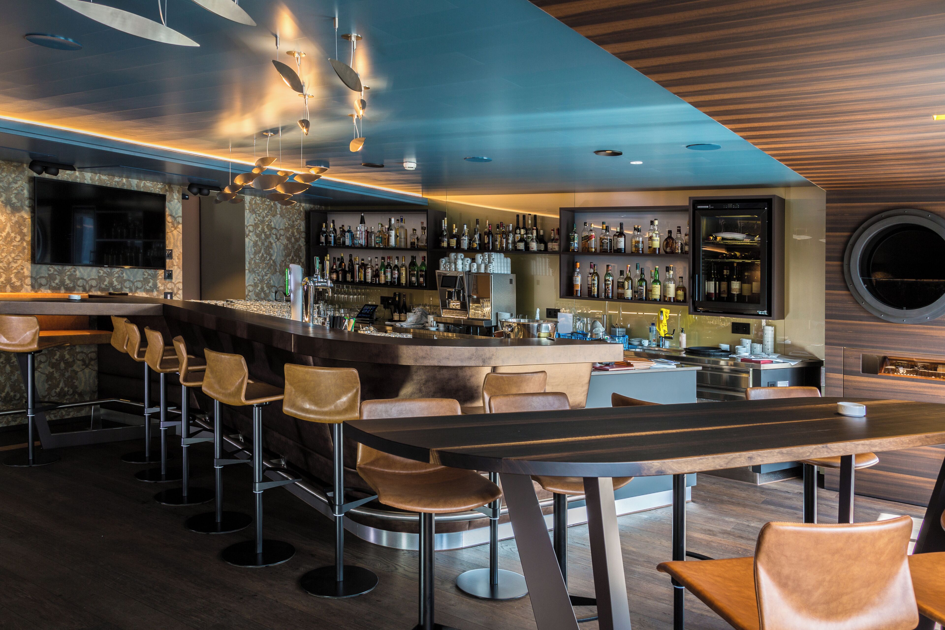 A modern bar area with a long counter, multiple stools, and a variety of bottles.