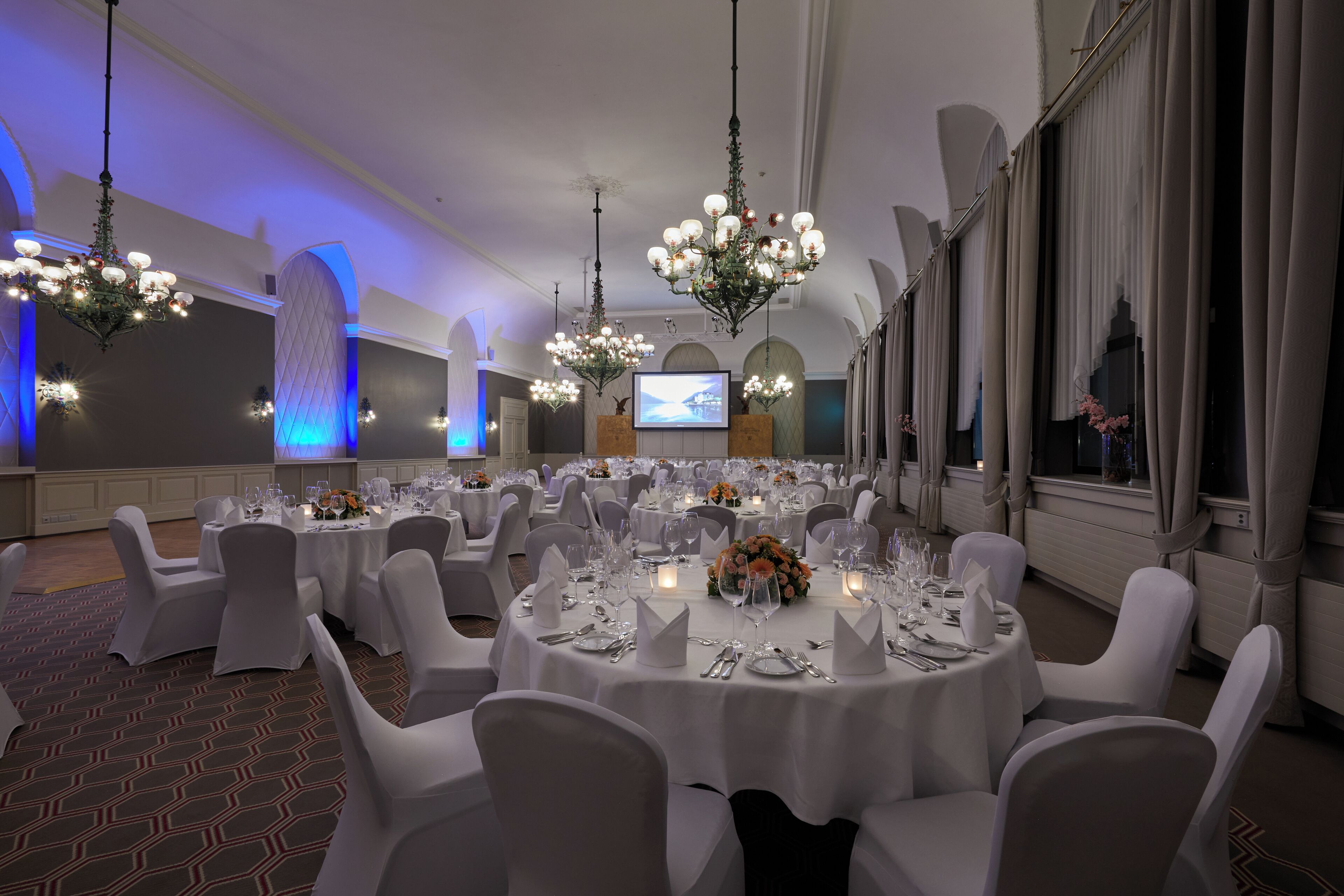 Un salón de banquetes con mesas redondas, sillas blancas, candelabros y una pantalla de proyección.