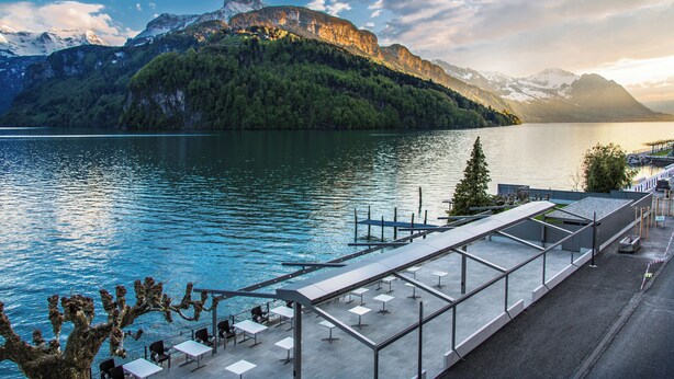 Eine Terrassenausrichtung am Seeufer mit Tischen und Stühlen, mit Blick auf einen See und Berge.