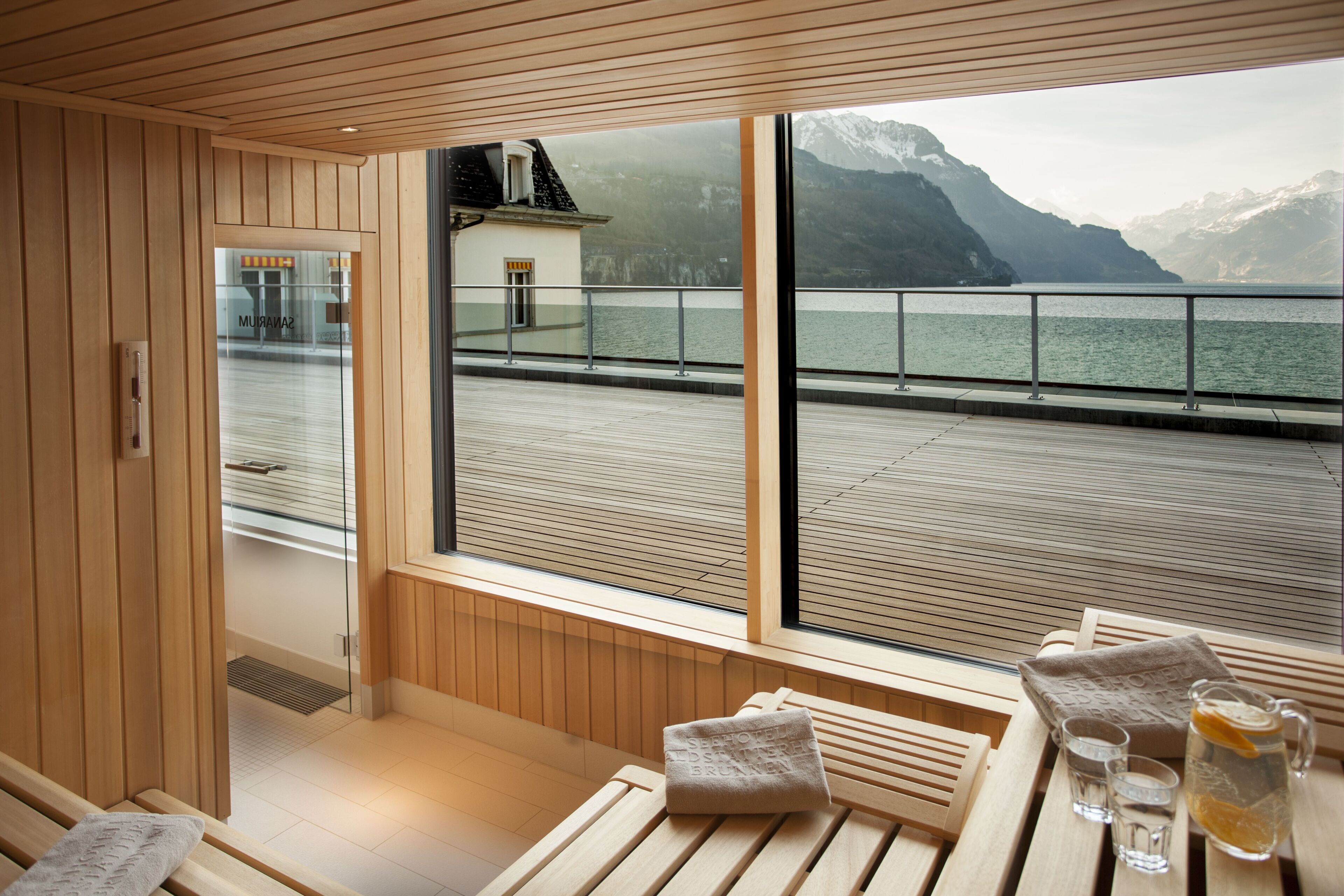 A wooden interior with a bench, a table with towels and glasses, and a view of a lake and mountains through large windows.