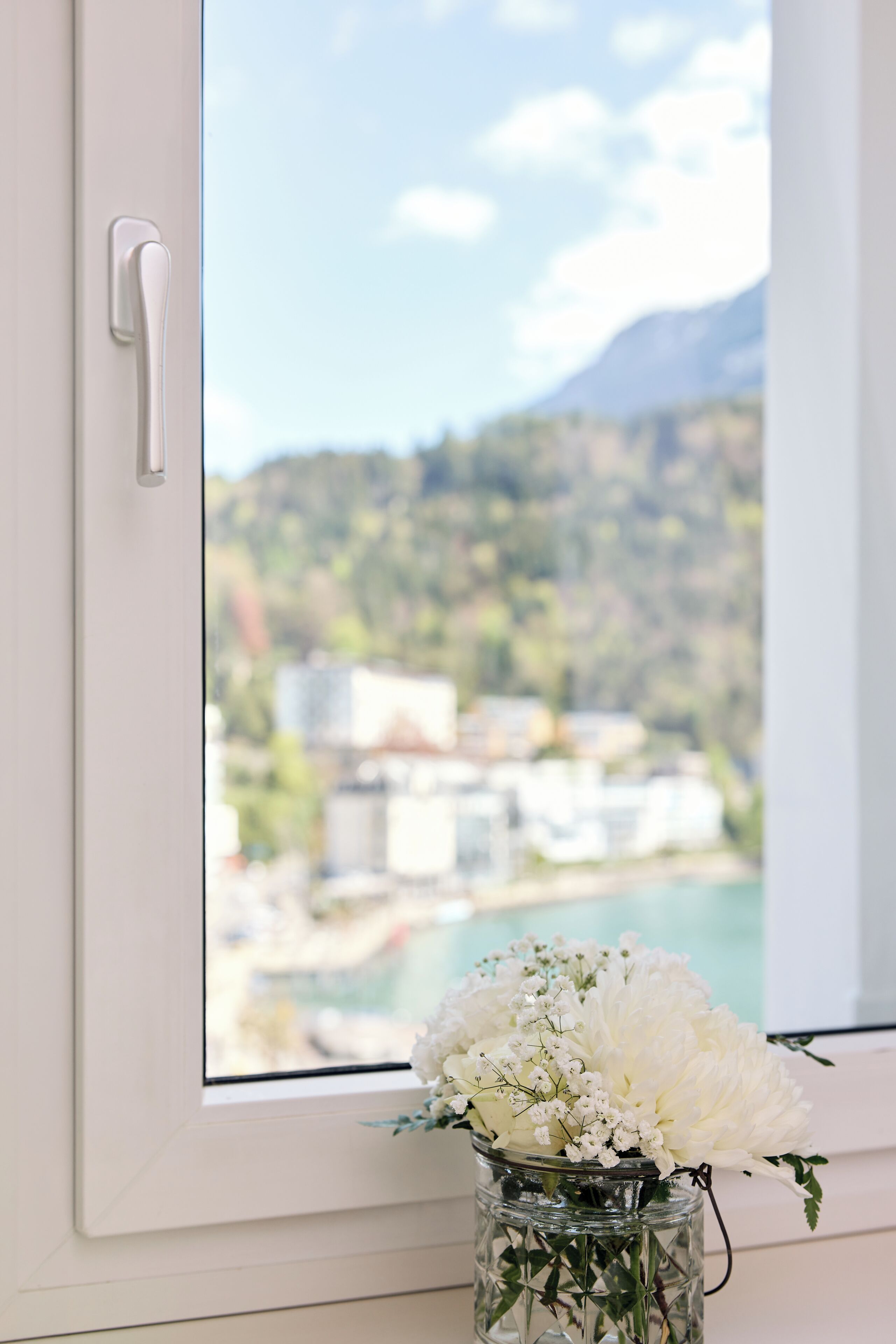 A window with a white door handle, a clear view of a mountainous landscape, and a vase with white flowers on a windowsill.