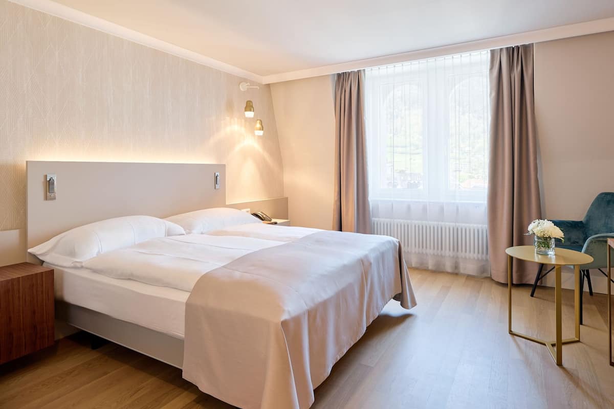 Une chambre d’hôtel comprenant un lit, une table de chevet, une lampe, une chaise, une petite table et une fenêtre avec des rideaux.