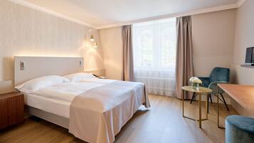 Une chambre d’hôtel comprenant un lit, une table de chevet, une lampe, une chaise, une petite table et une fenêtre avec des rideaux.
