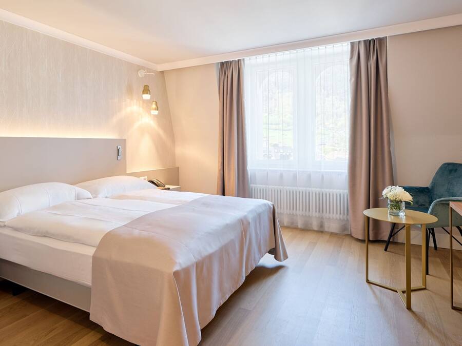 Une chambre d’hôtel comprenant un lit, une table de chevet, une lampe, une chaise, une petite table et une fenêtre avec des rideaux.