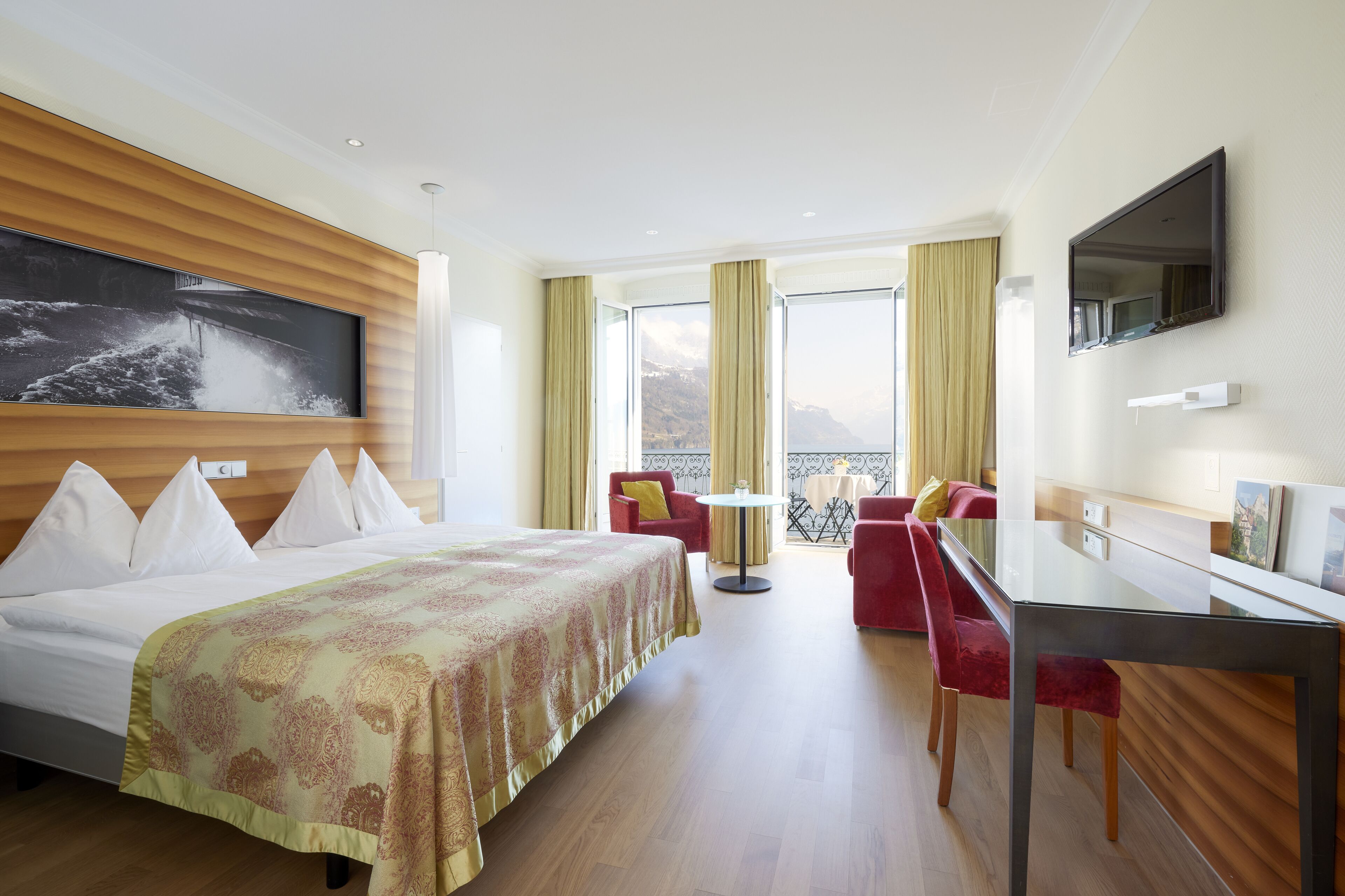 A hotel room with a bed, a TV, a dining table, and a balcony with red chairs.