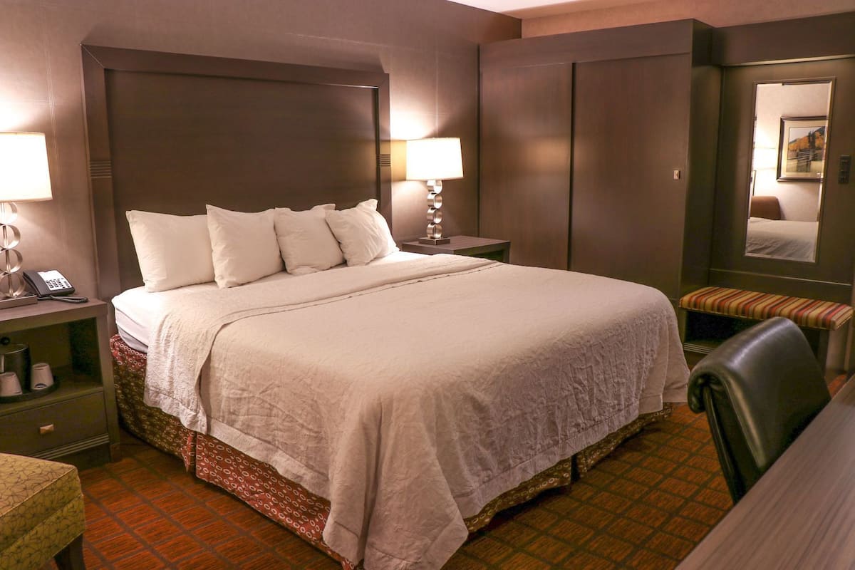A hotel room with a large bed, bedside lamps, a nightstand, a chair, a desk, and a mirror.