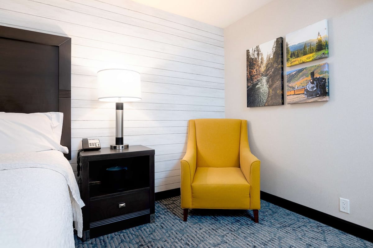 A hotel room with a bed, a nightstand with a lamp, a yellow armchair, and two landscape paintings on the wall.