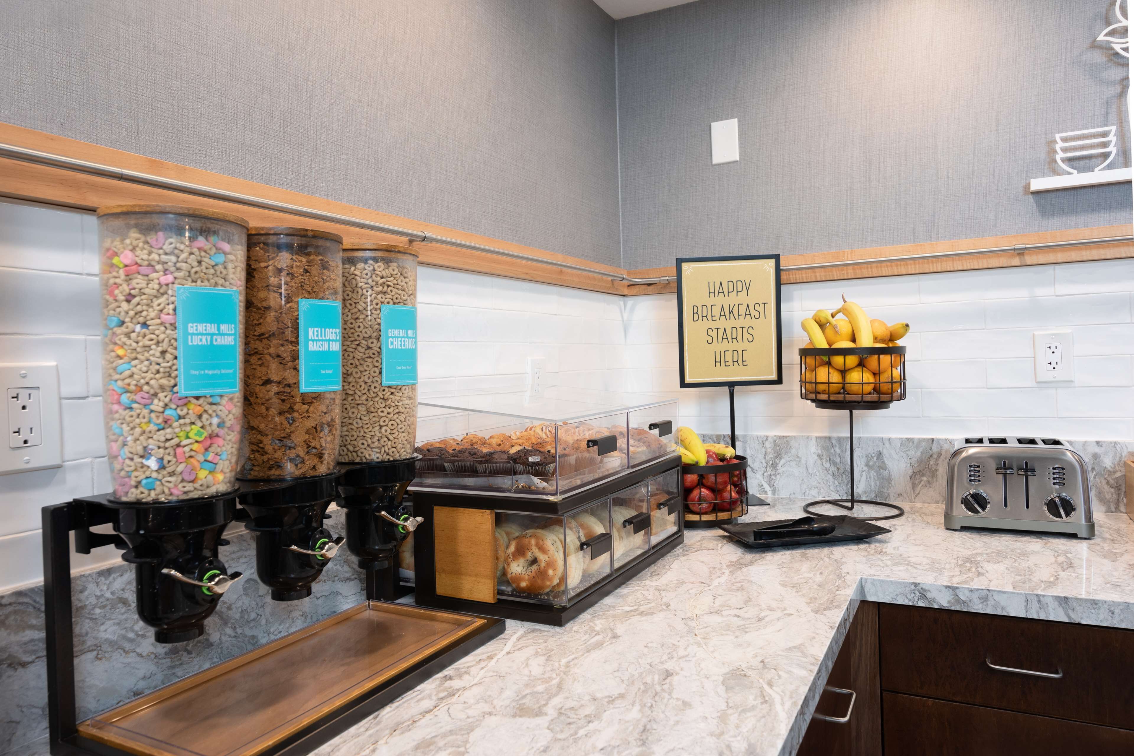 A breakfast station with cereal dispensers, a sign indicating breakfast starts here, a fruit basket, and a toaster.
