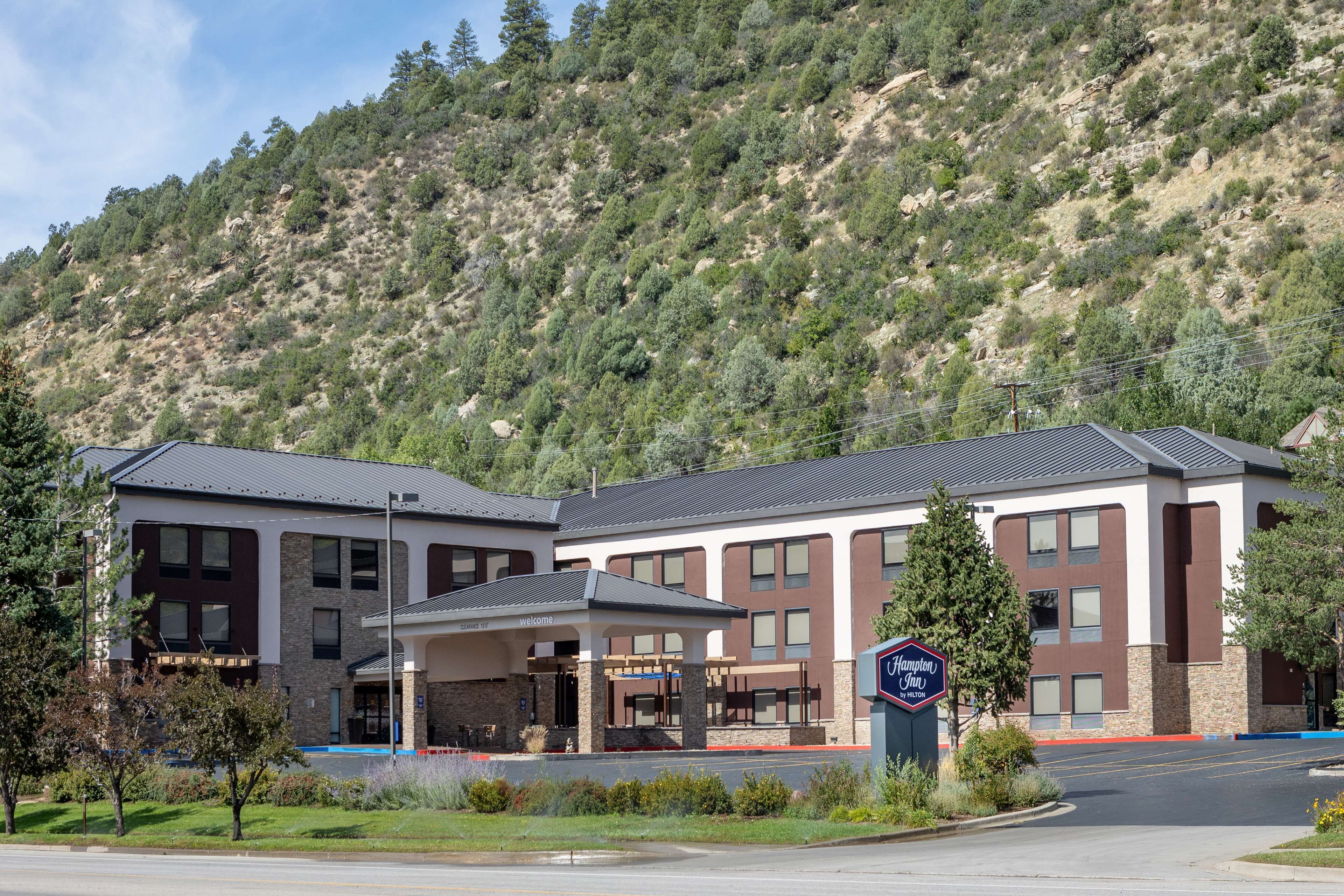 A modern hotel with a covered entrance, a sign indicating it is a Hampton Inn, and a mountainous backdrop.