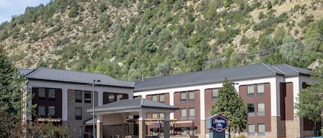 A modern hotel with a covered entrance, a sign indicating it is a Hampton Inn, and a mountainous backdrop.