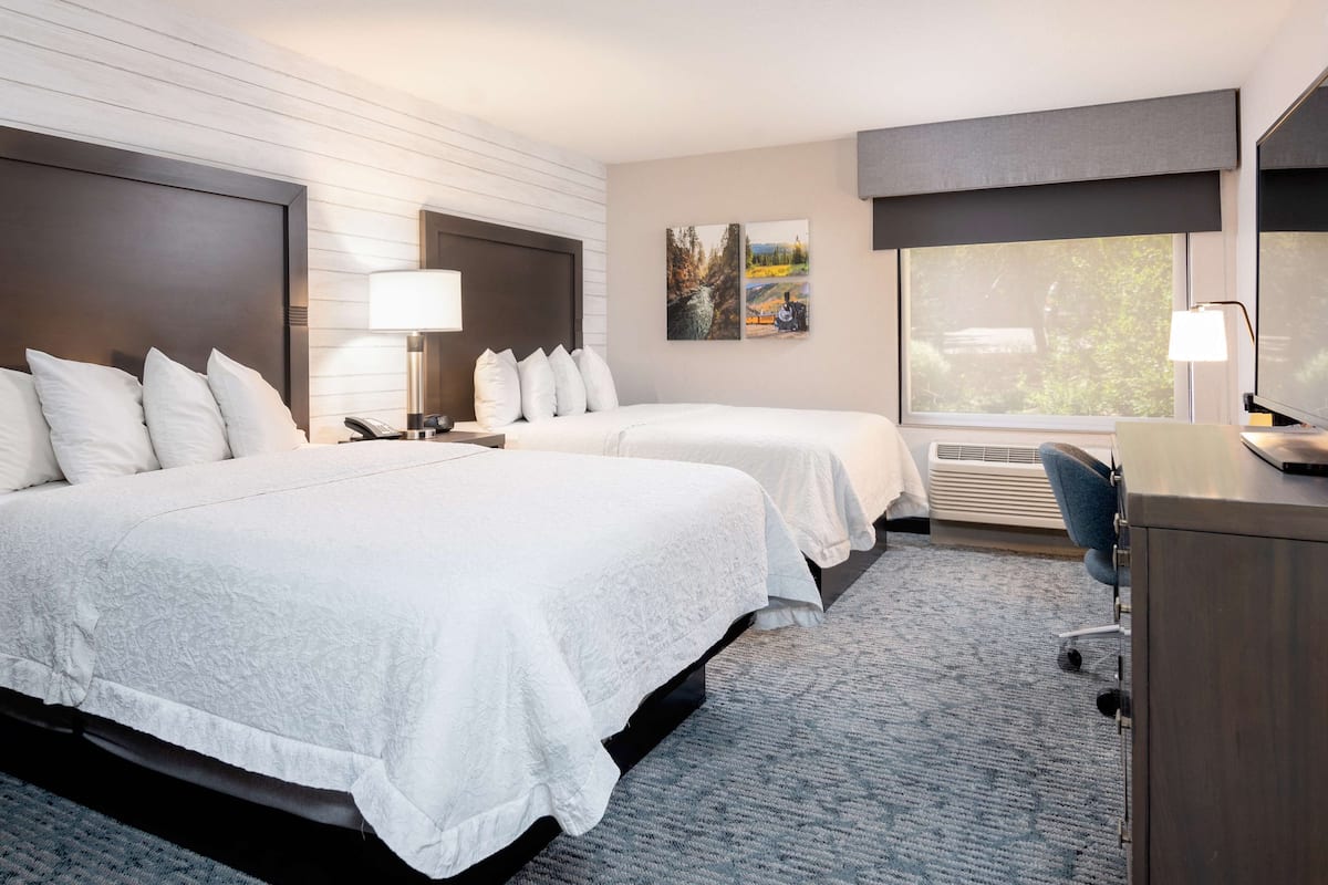 A hotel room with two beds, a desk, a chair, a lamp, and a window with a view of trees.