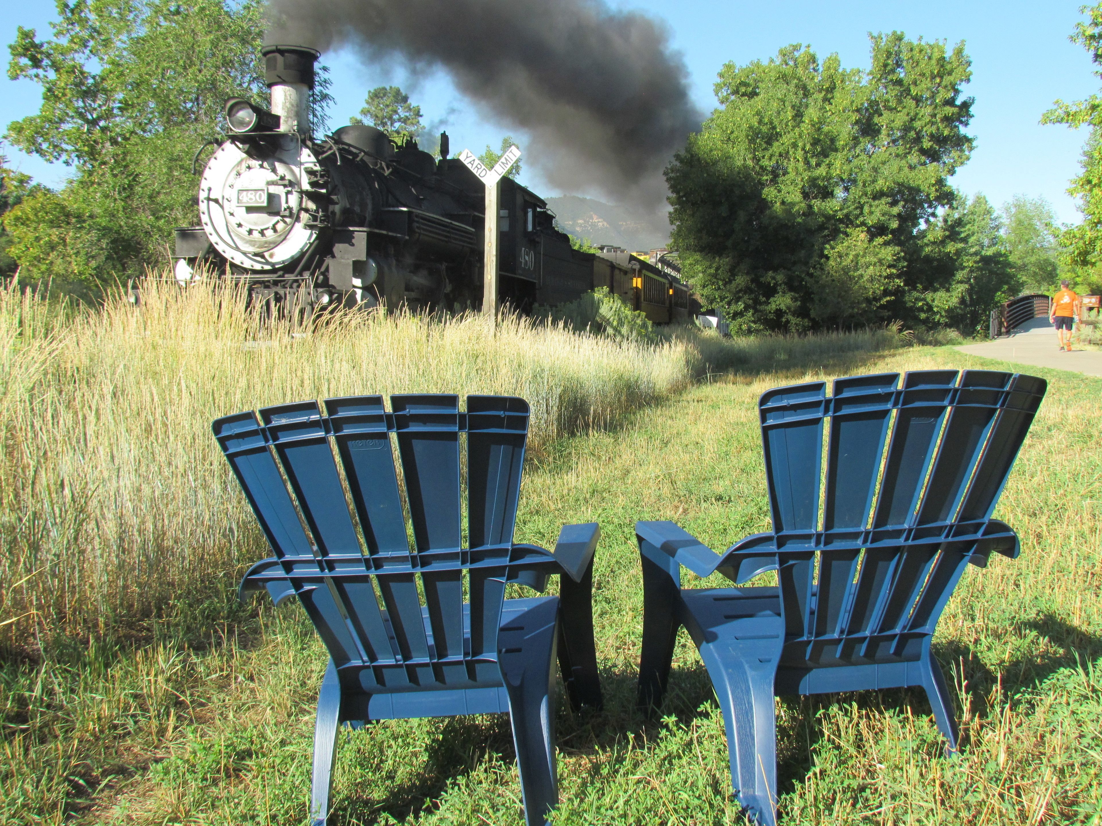 Two blue Adirondack chairs in a grassy area with a steam locomotive in the background.
