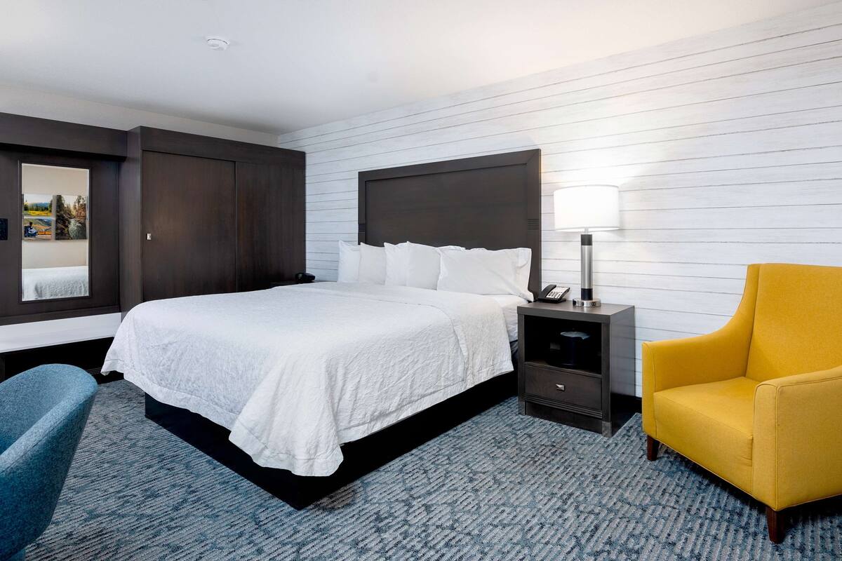 A hotel room with a large bed, a bedside table with a lamp, a chair, and a view of the outdoors through a window.