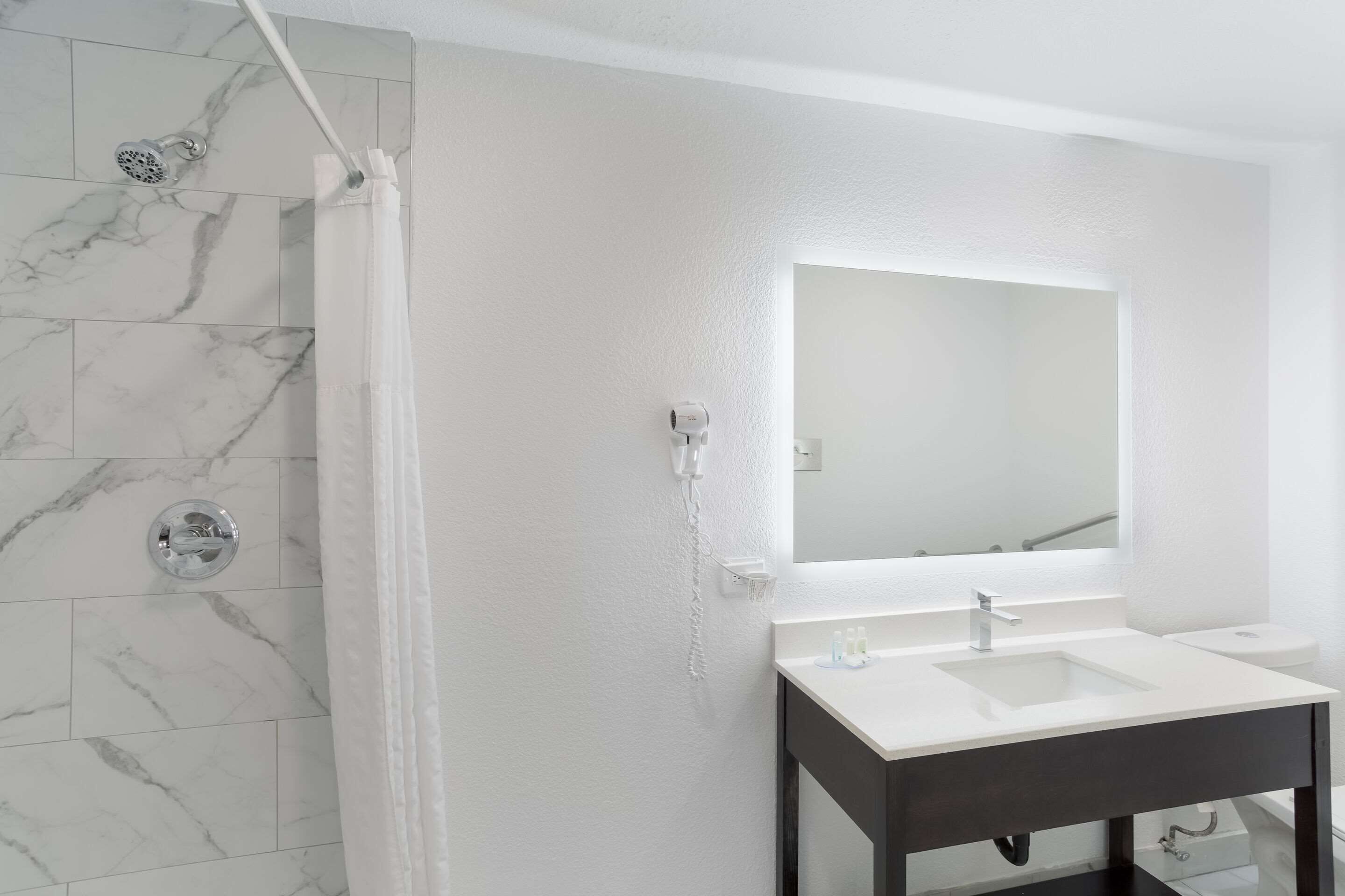 A bathroom with a white marble shower, a white sink with a dark vanity, and a large mirror above the sink.