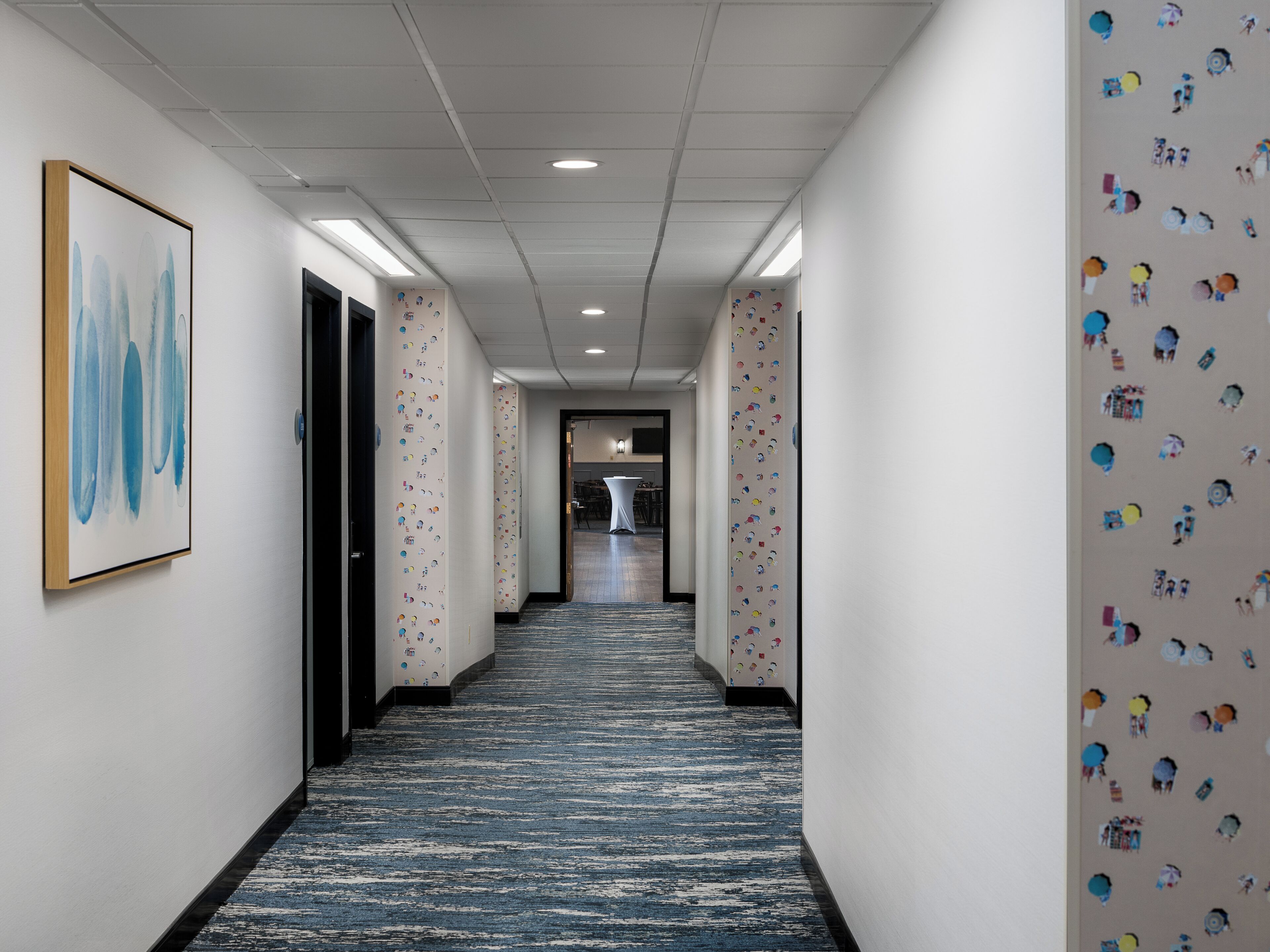 Un pasillo de hotel con alfombra estampada, paredes blancas y una obra de arte enmarcada a la izquierda.