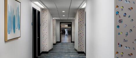 Un pasillo de hotel con alfombra estampada, paredes blancas y una obra de arte enmarcada a la izquierda.