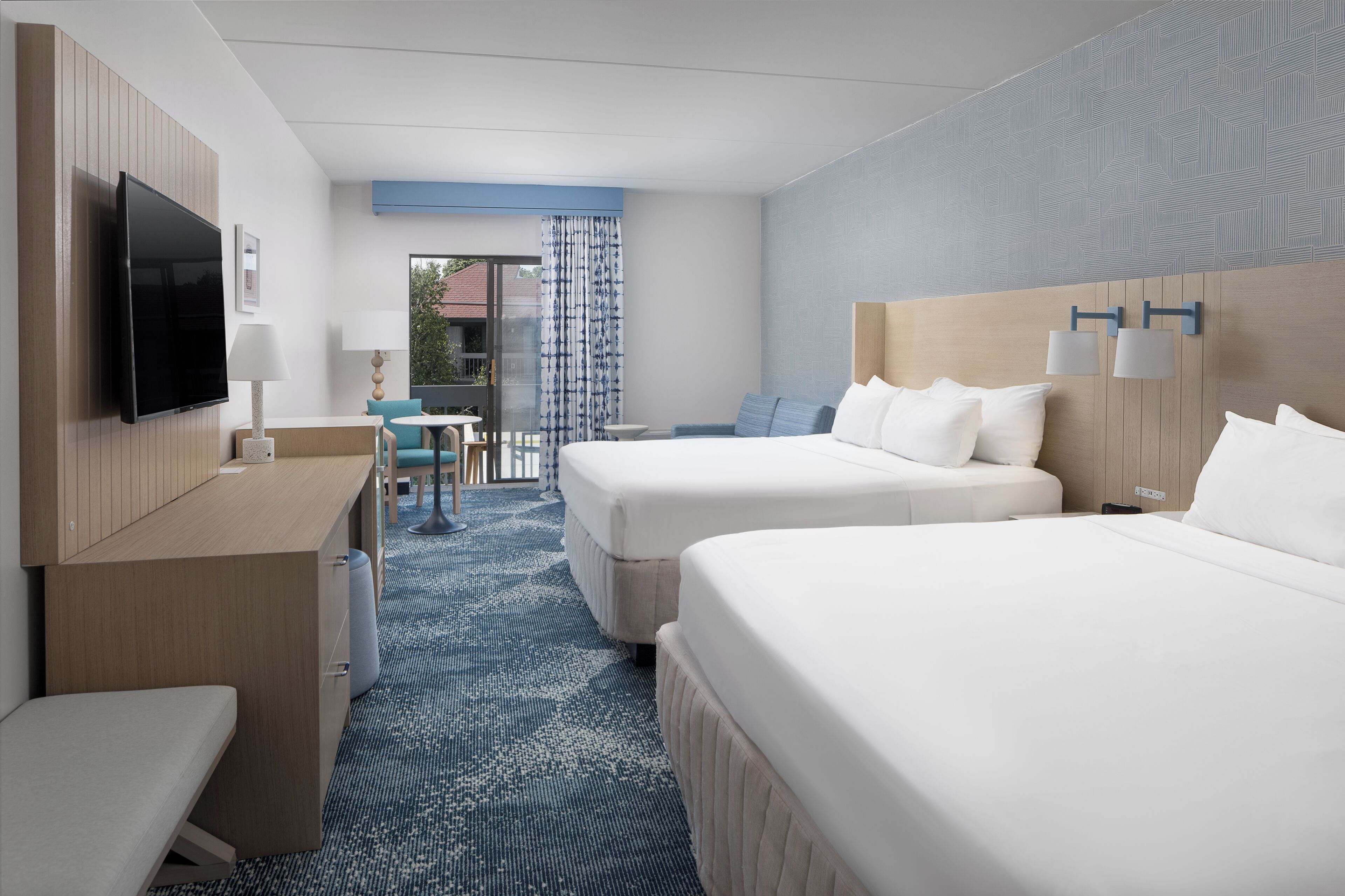 A hotel room with two beds, a desk, a TV, and a view of the outdoors.