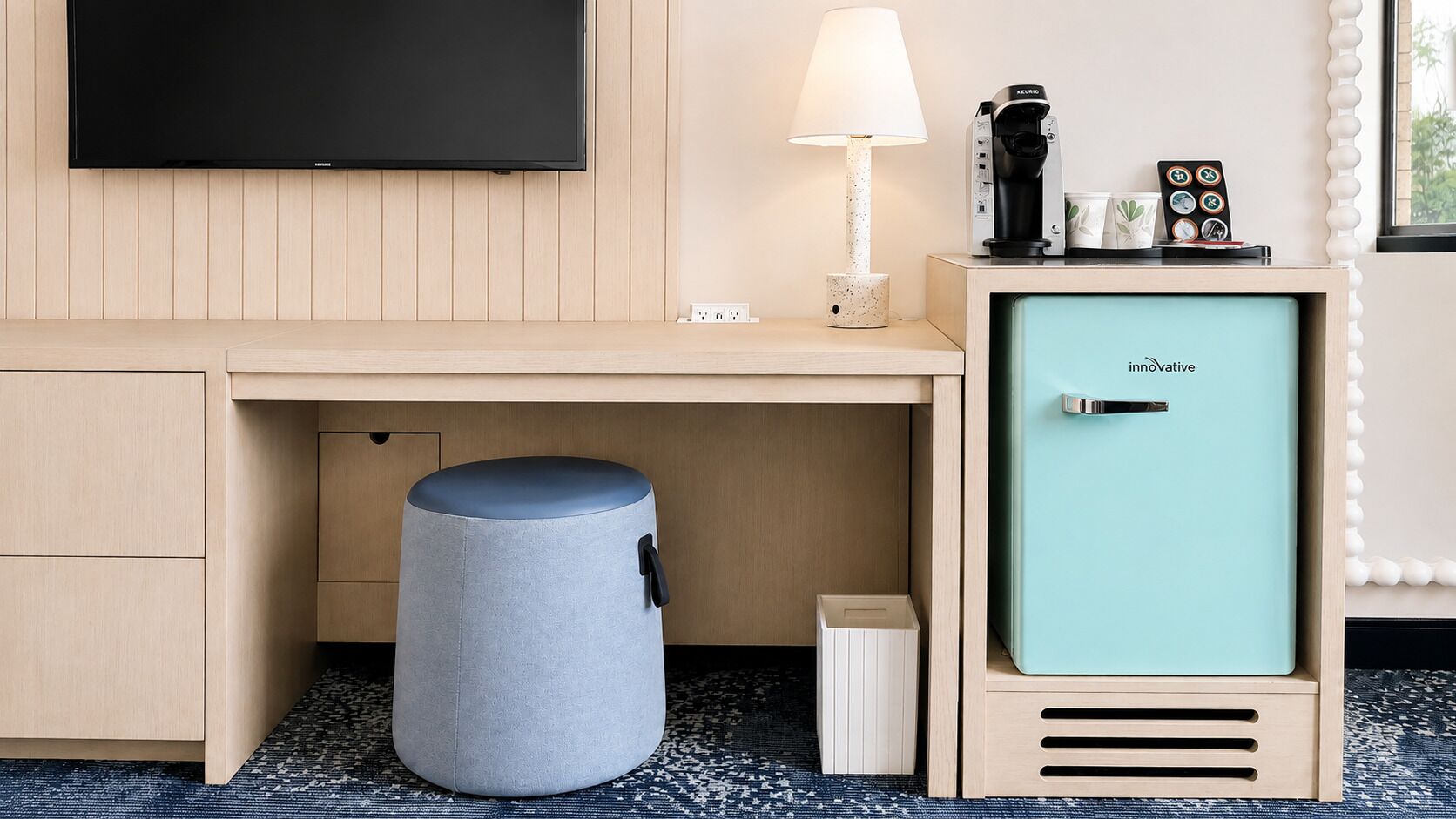 A hotel room with a flat-screen TV, a lamp, a coffee machine, a mini fridge, and a trash bin.
