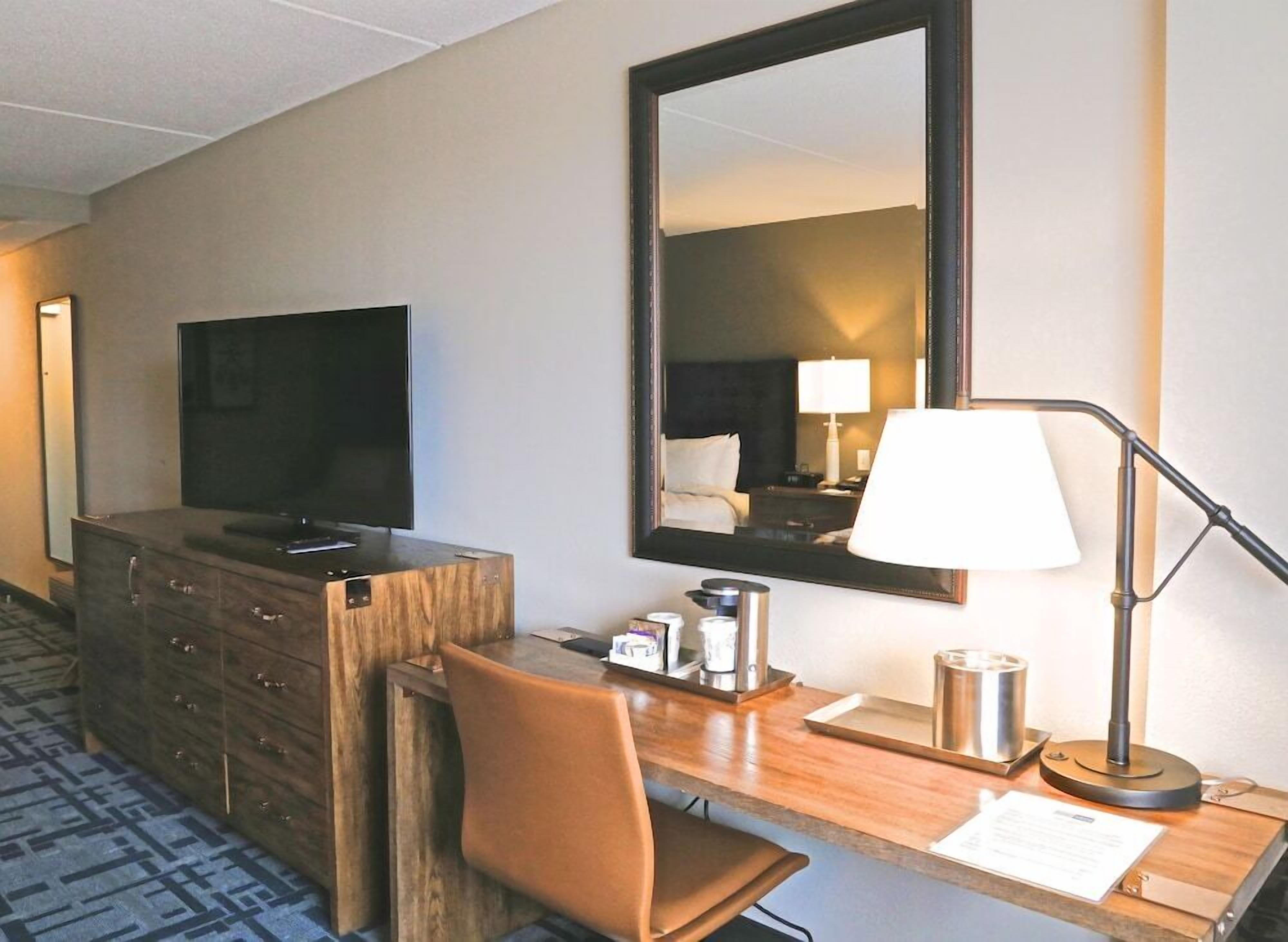 Una habitación de hotel con un escritorio de madera, televisión, espejo, lámpara y una silla.