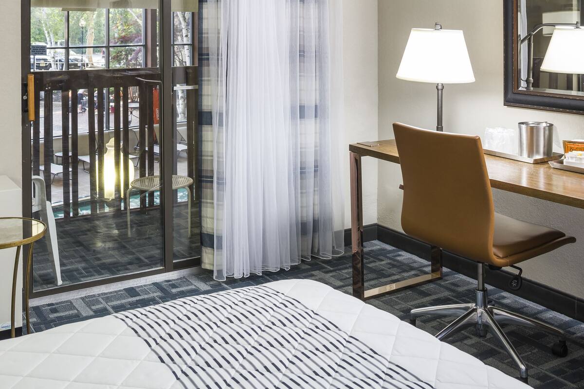 Ein Hotelzimmer mit einem Bett, einem Schreibtisch mit Lampe, einem Sessel und Blick durch eine Glastür.