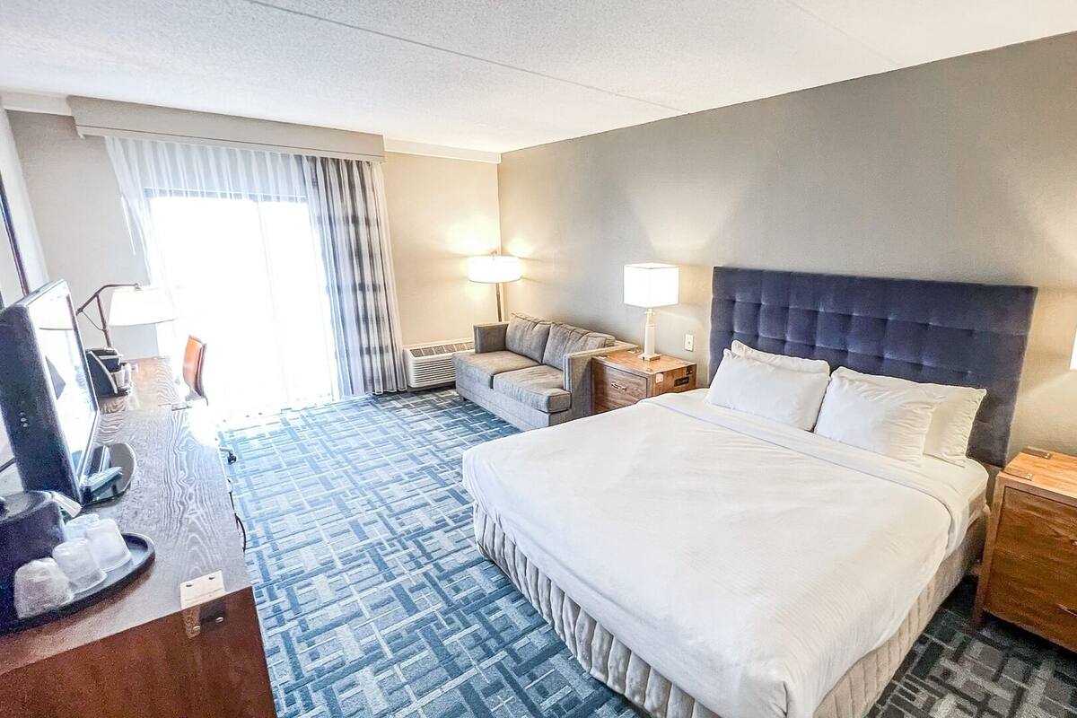 Ein Hotelzimmer mit einem großen Bett, zwei Nachttischen inklusive Lampen, einem Sofa, einem Schreibtisch mit Computer sowie einem Fenster mit Vorhängen.