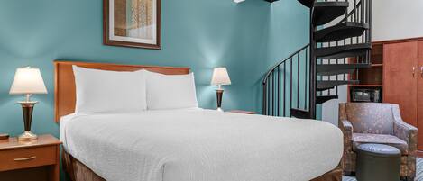A hotel room with a large bed, two bedside tables with lamps, a chair, a small ottoman, a framed artwork on the wall, and a spiral staircase.