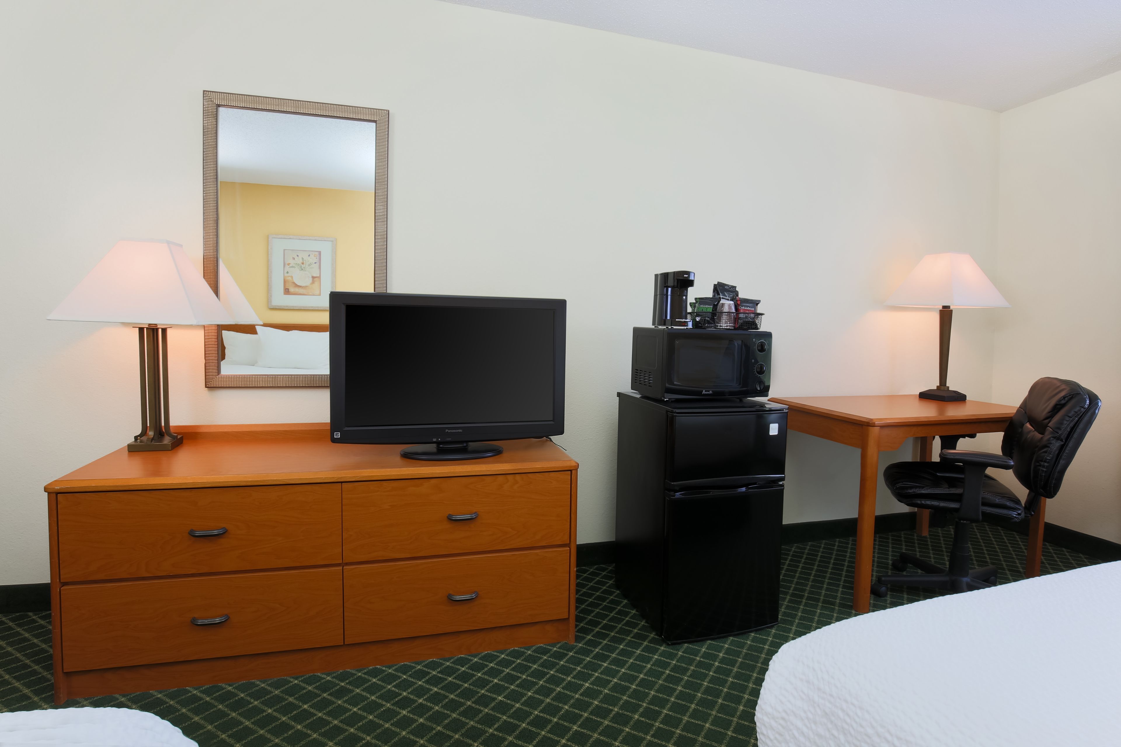 A hotel room with a bed, a desk with a chair, a television, a refrigerator, and two lamps.