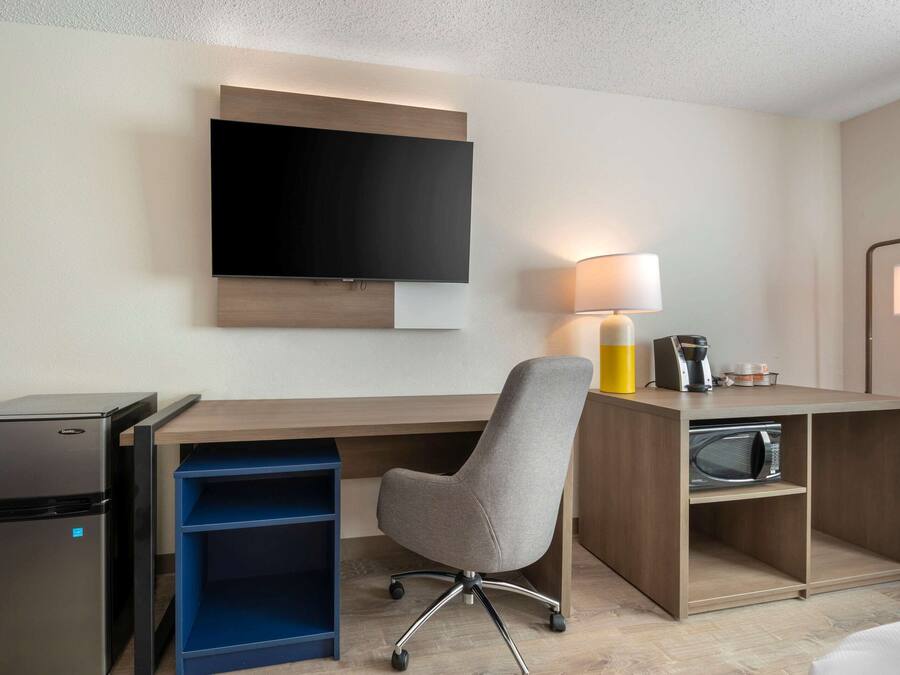 A hotel room with a flat-screen TV mounted on the wall, a desk with a lamp, a microwave, and a small refrigerator.