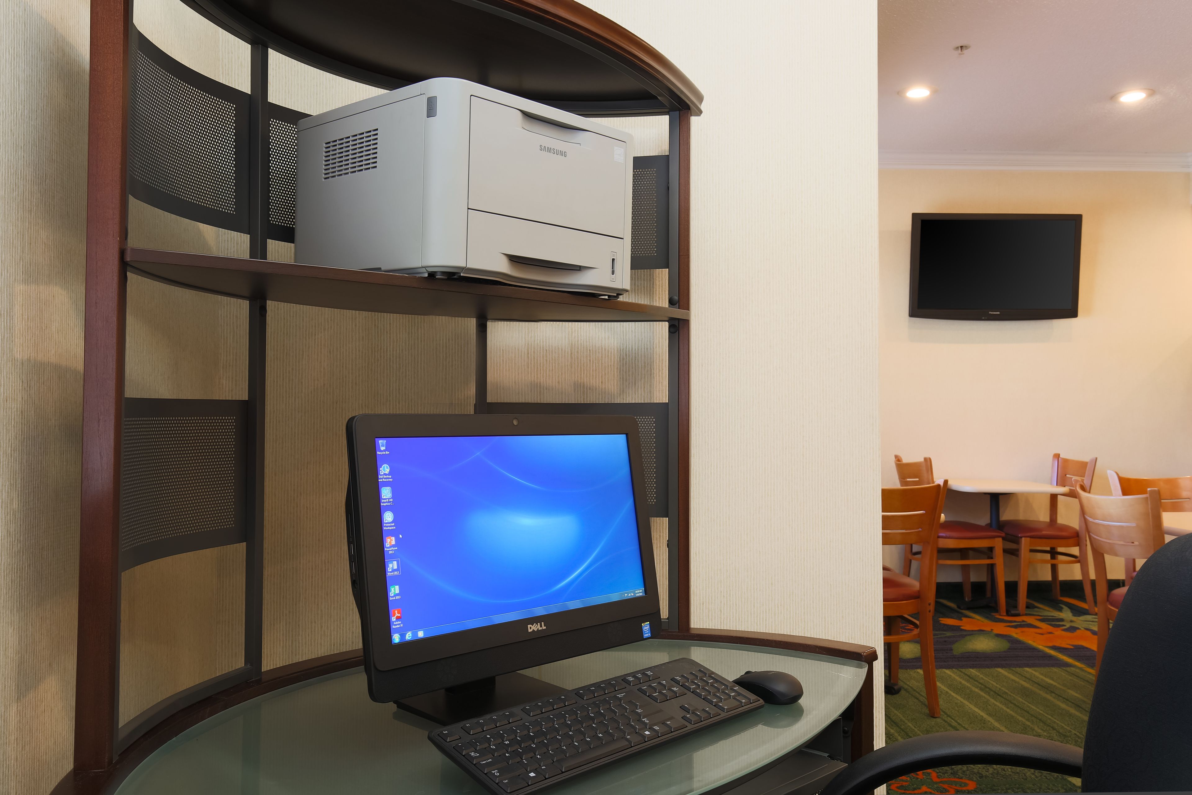 A hotel room with a desk, computer, printer, and a TV mounted on the wall.