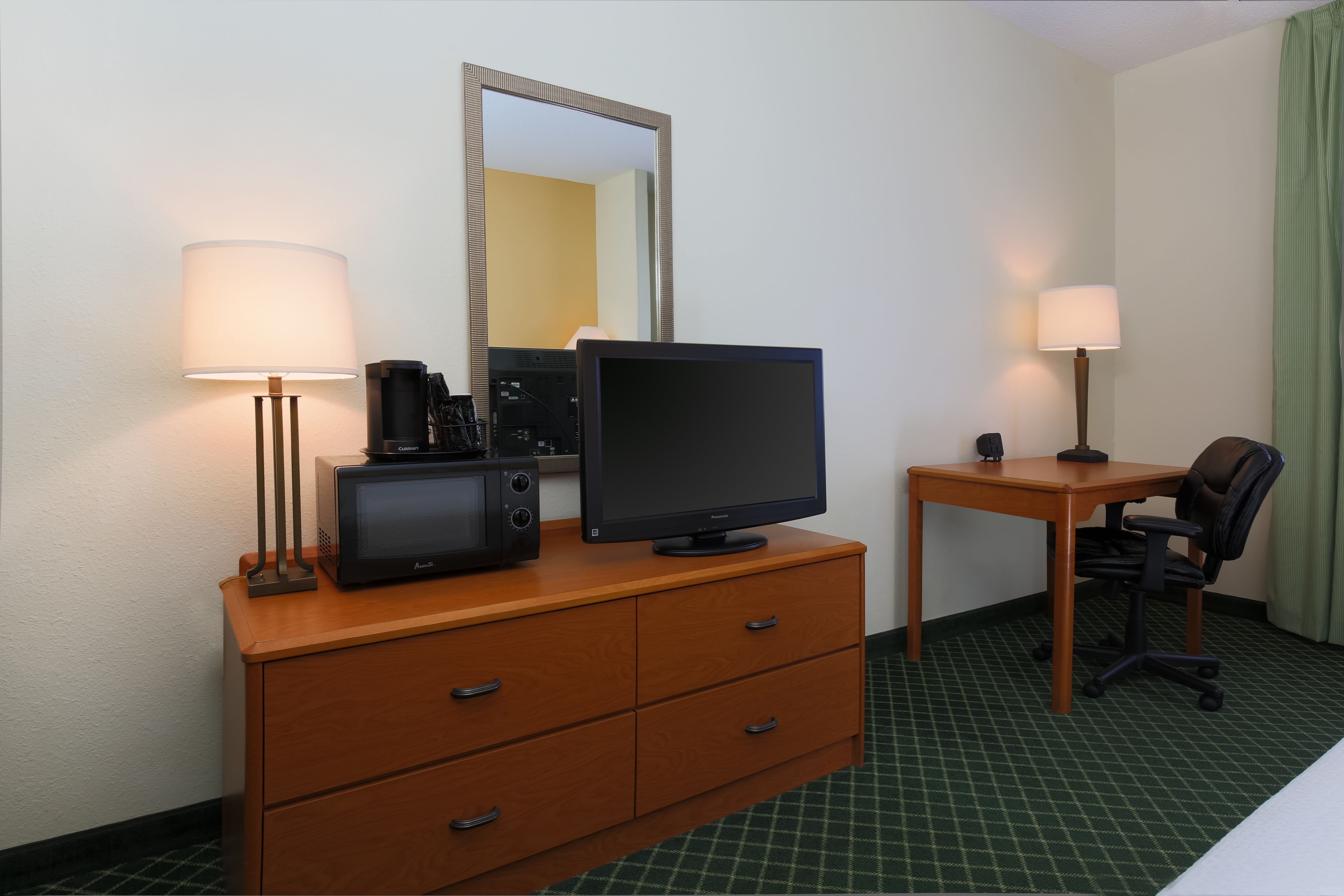 A hotel room with a wooden desk, a television, a lamp, a chair, and a dresser.
