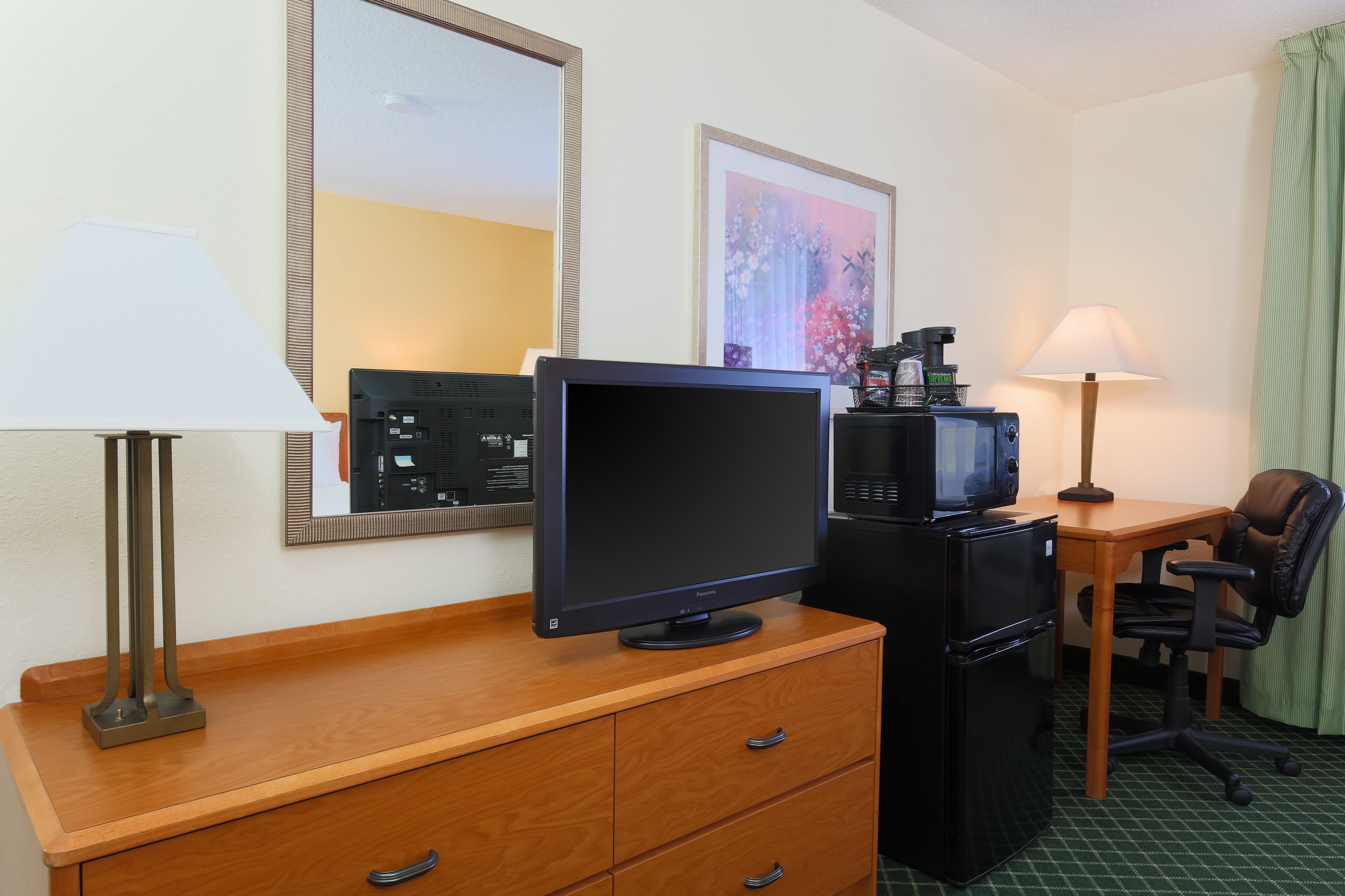 A hotel room with a wooden desk, a flat-screen TV, a refrigerator, a lamp, a chair, and a framed picture on the wall.