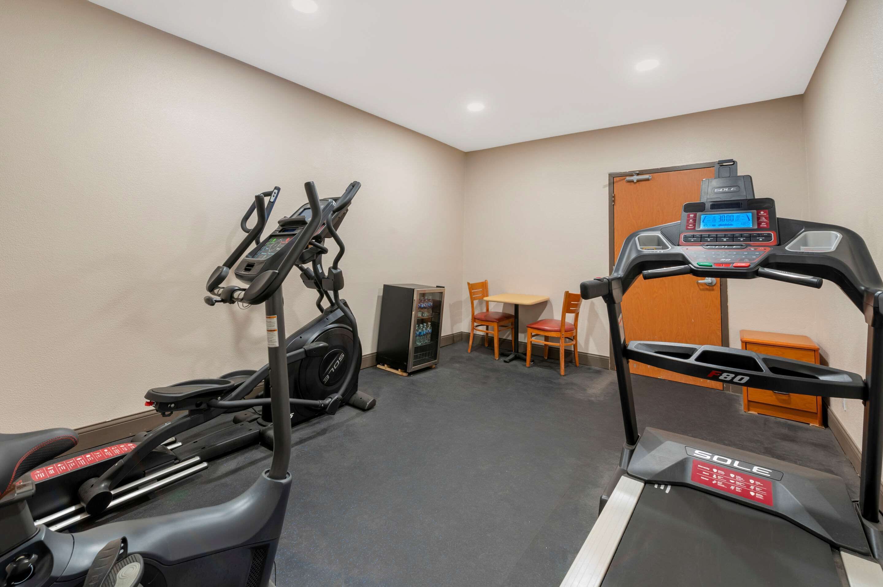 Un gimnasio con cintas de correr, elíptica y bicicleta estática. Hay una pequeña mesa y sillas, y una puerta al fondo.