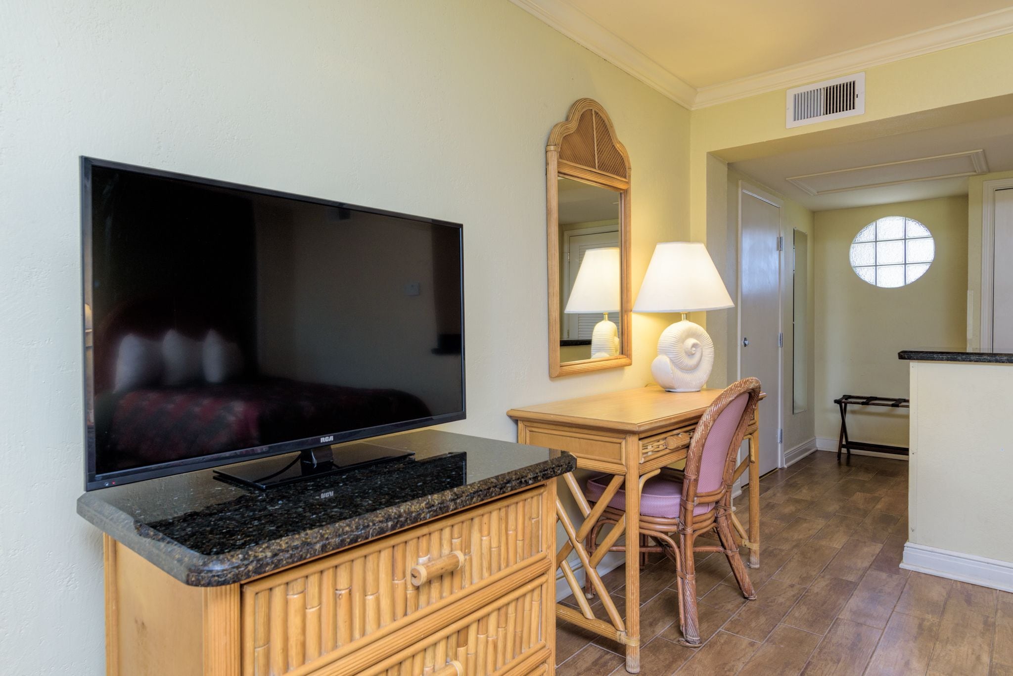 Une chambre d’hôtel équipée d’un téléviseur à écran plat, d’un bureau en bois avec une lampe, d’un fauteuil en osier et d’un grand miroir.